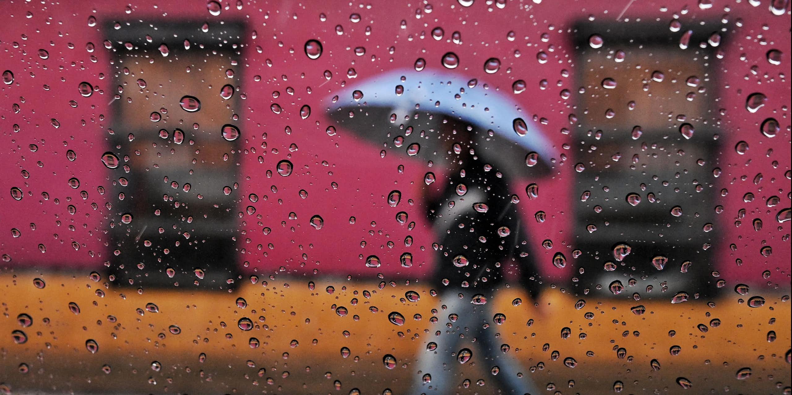 A person walks in the rain past brightly colored buildings with an umbrella. The photo is shot through a window with raindrops on the glass.