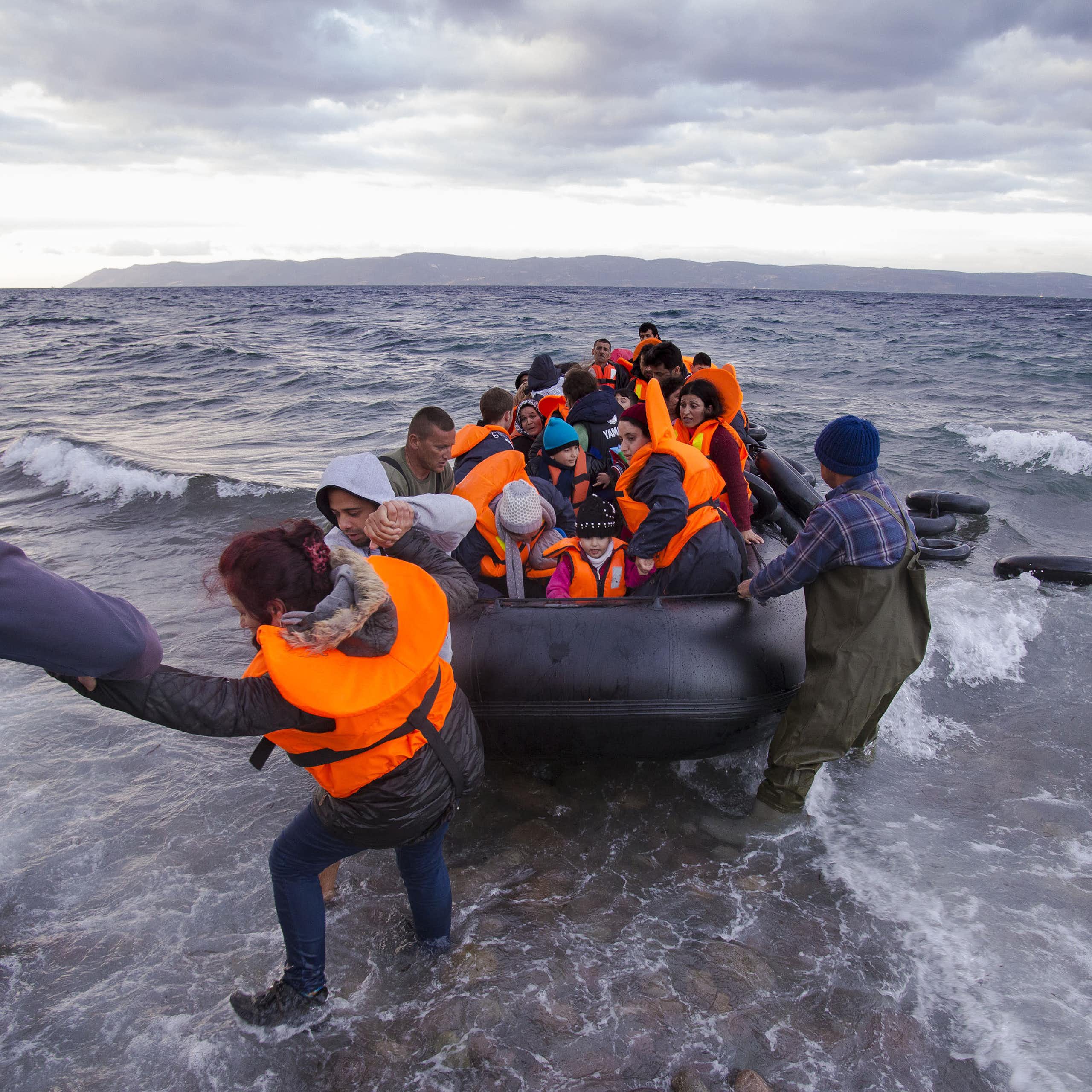 People on shore help pull a rubber dinghy full of people in life jackets from the ocean