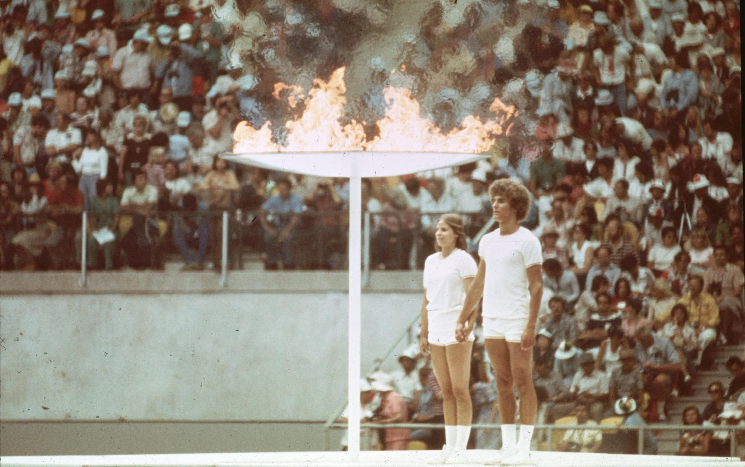 A vintage colour photo of two young people - a man and woman - standing at attention next to a giant torch that is flaming. In the background, packed stands of a stadium.