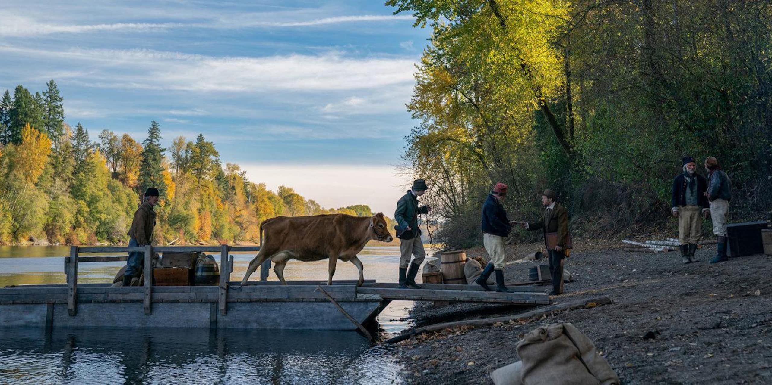 Four men lead a cow along a wooden jetty on the shore of a river surrounded by trees 