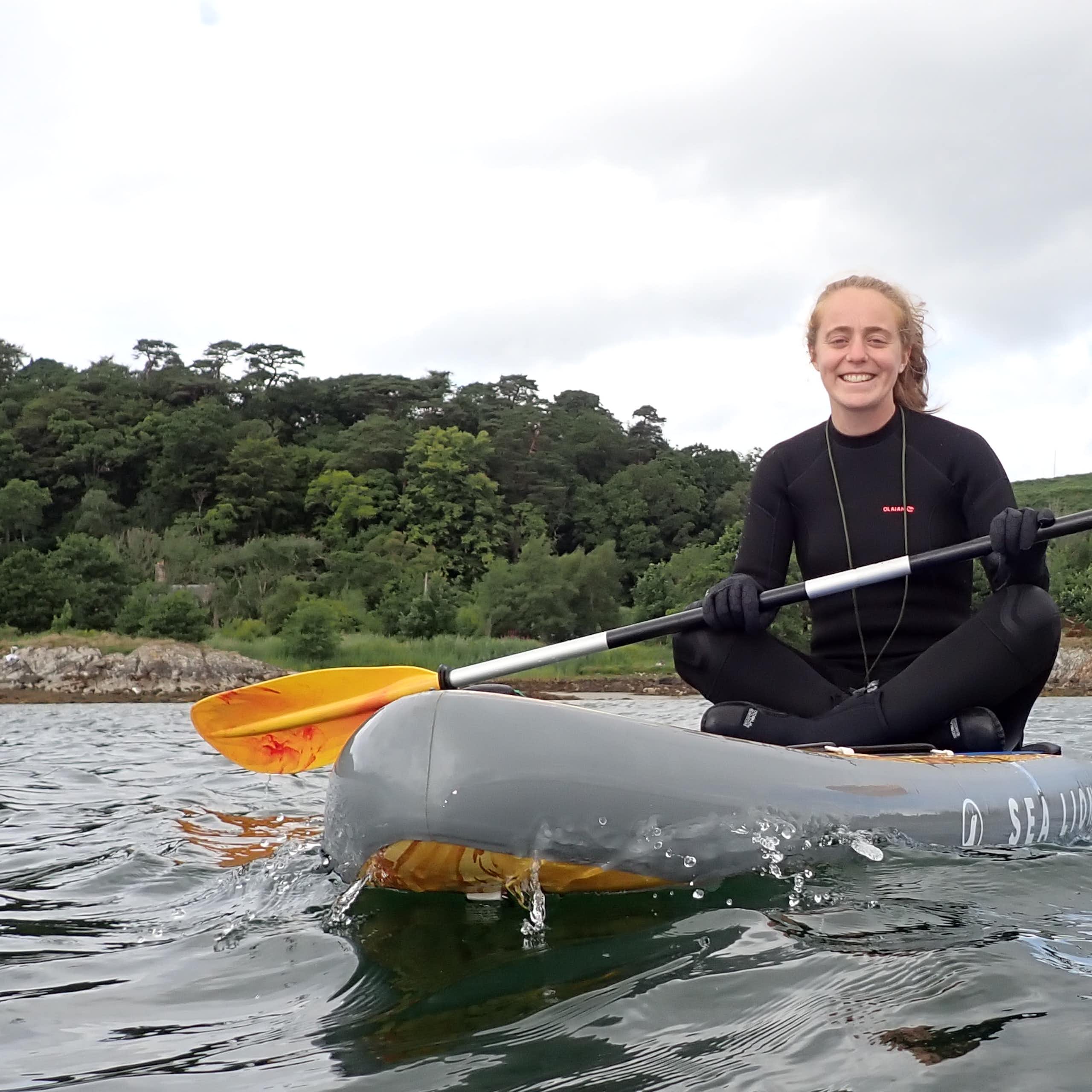 woman in black wetsuit sitting on kayak on sea, trees in background, white sky above