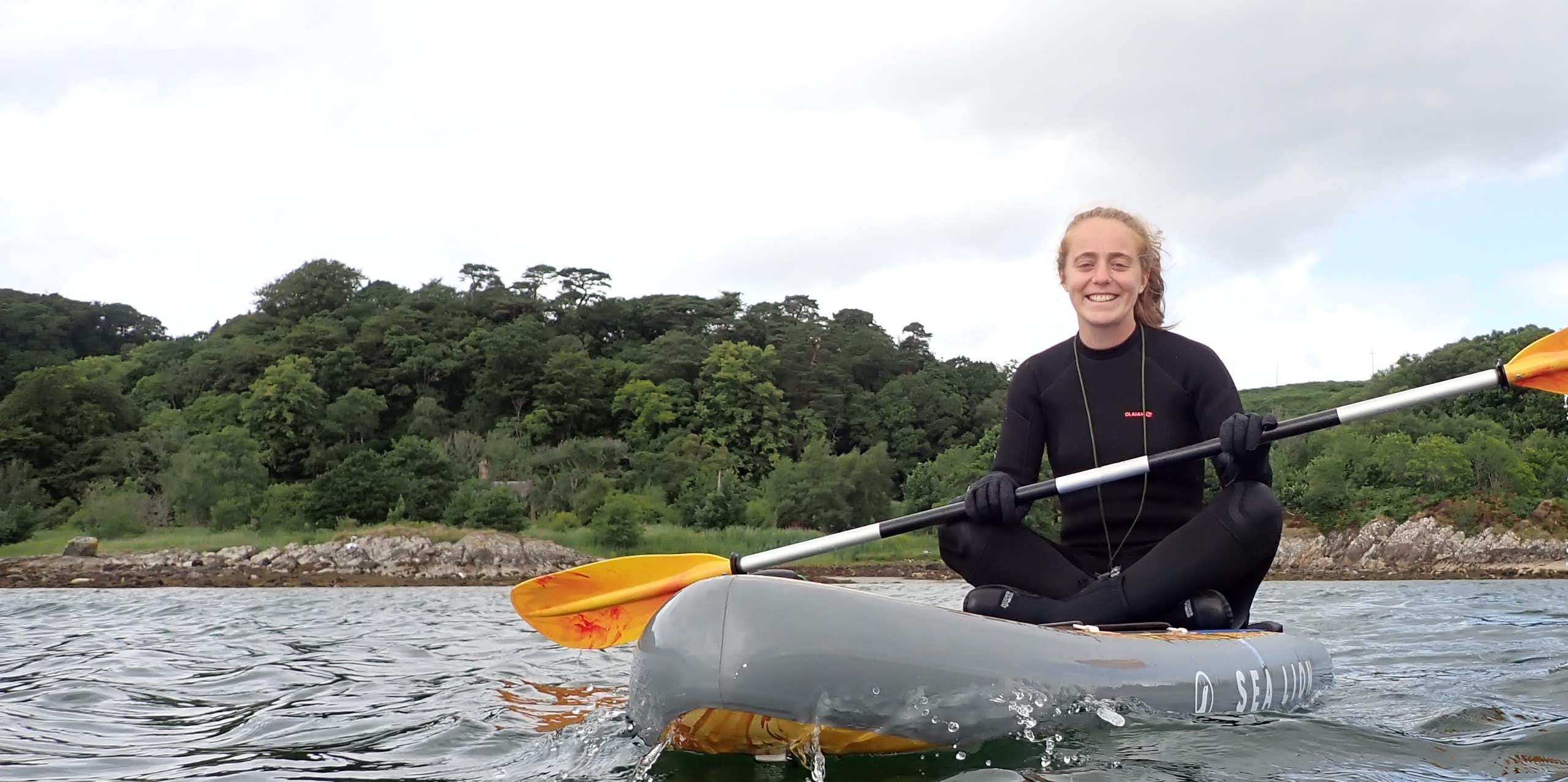 woman in black wetsuit sitting on kayak on sea, trees in background, white sky above