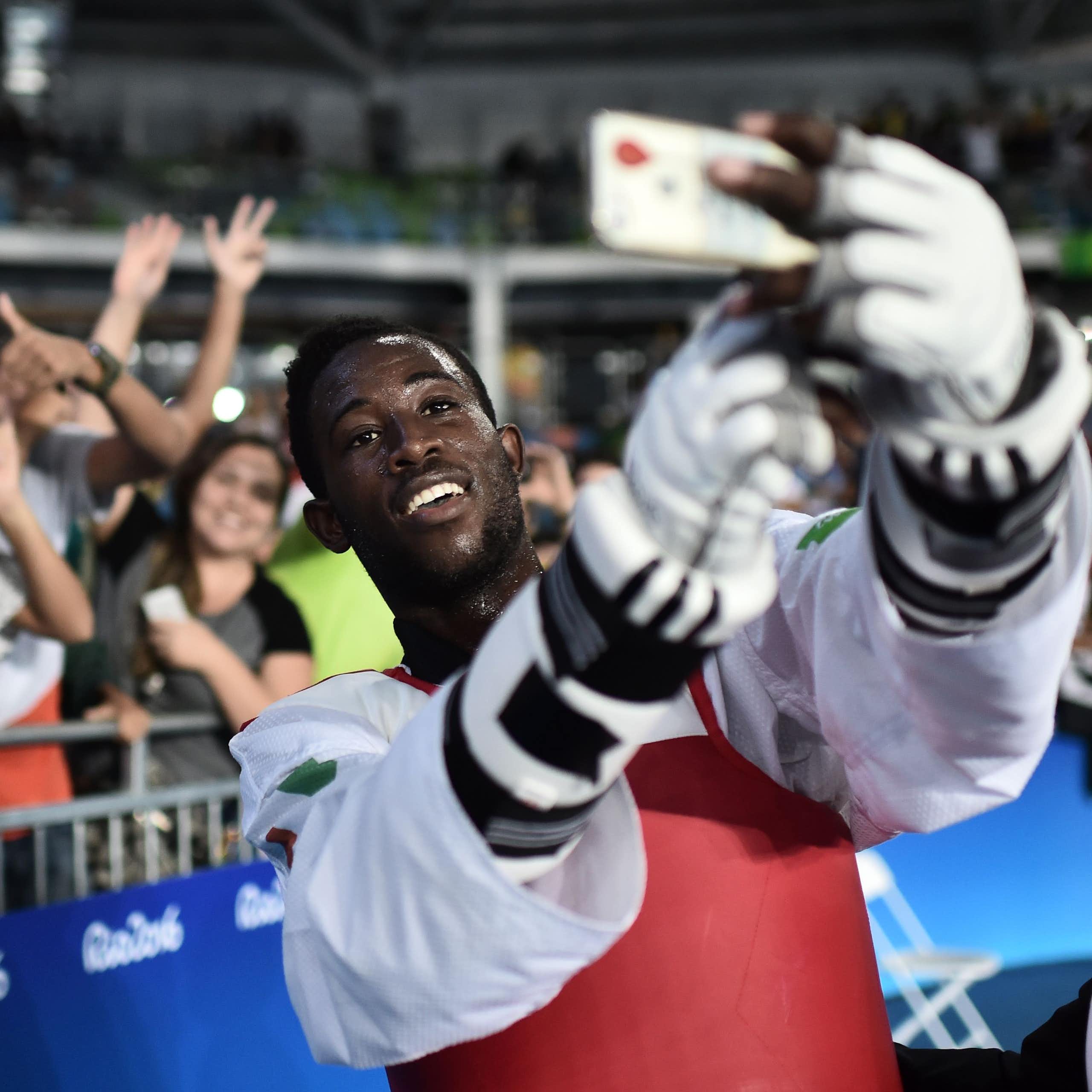 A grinning athlete in protective gloves and chest guard holds up a phone to take photos with a group of grinning people