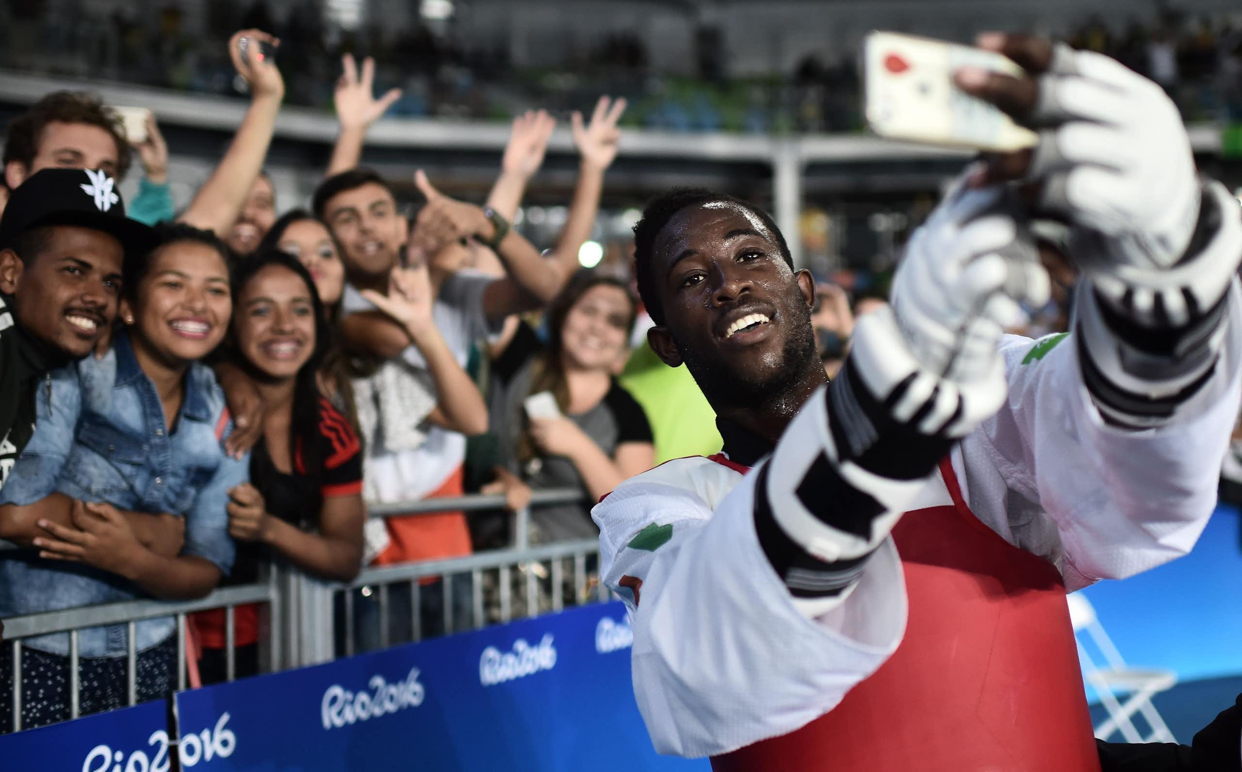 A grinning athlete in protective gloves and chest guard holds up a phone to take photos with a group of grinning people