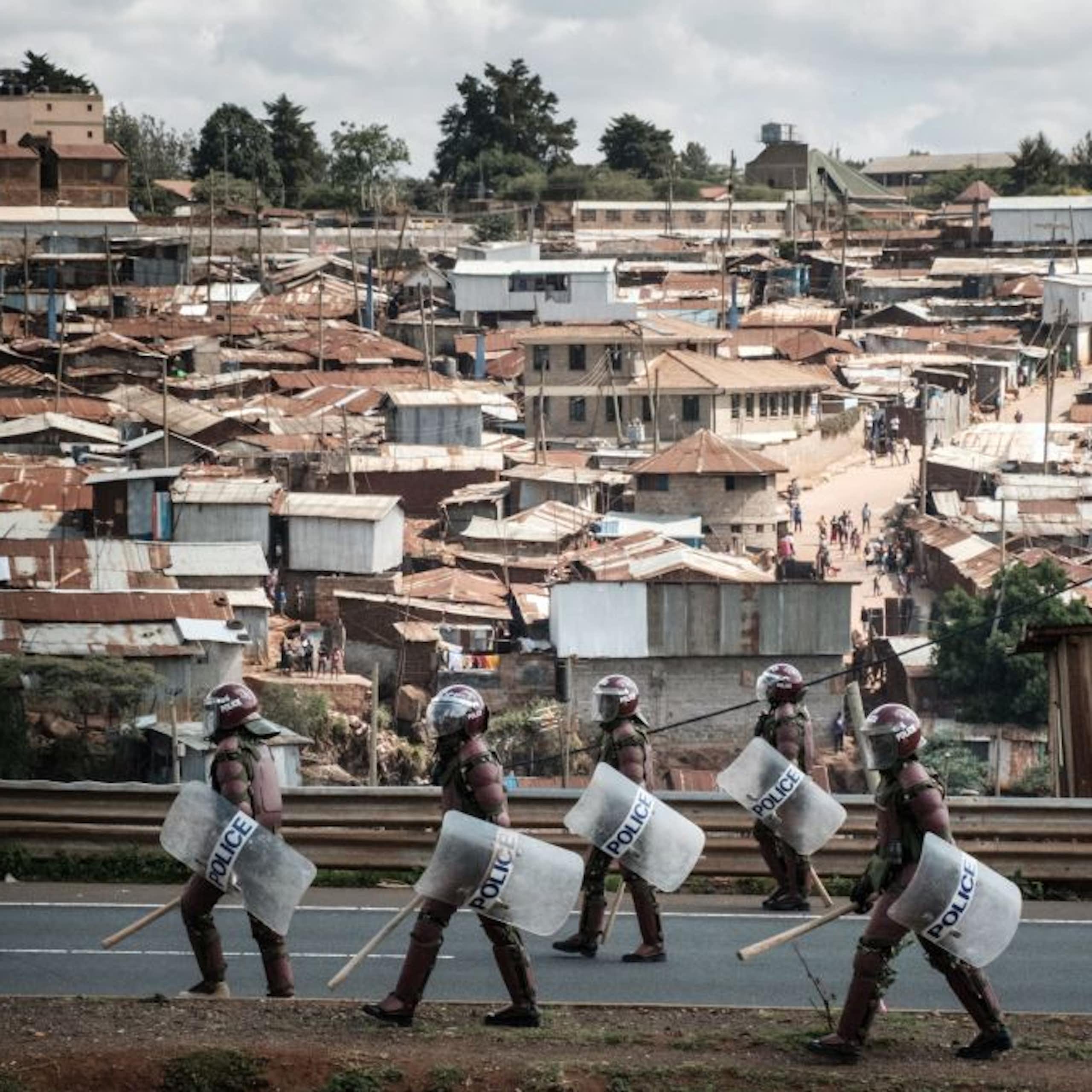 People in helmets, carrying truncheons and shields, walk on a road with a dense informal settlement in the background