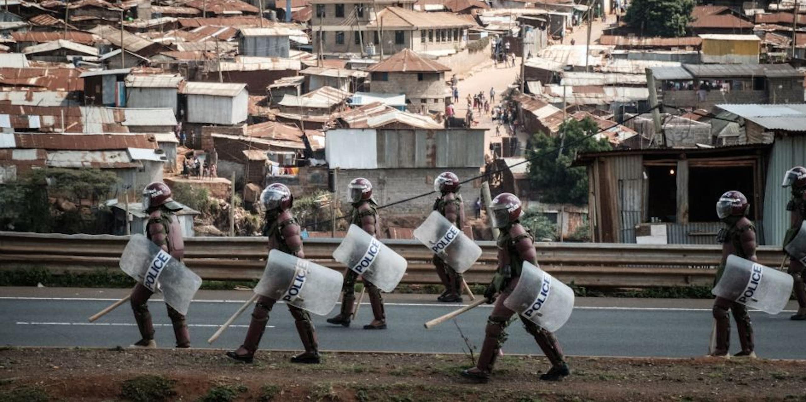 People in helmets, carrying truncheons and shields, walk on a road with a dense informal settlement in the background