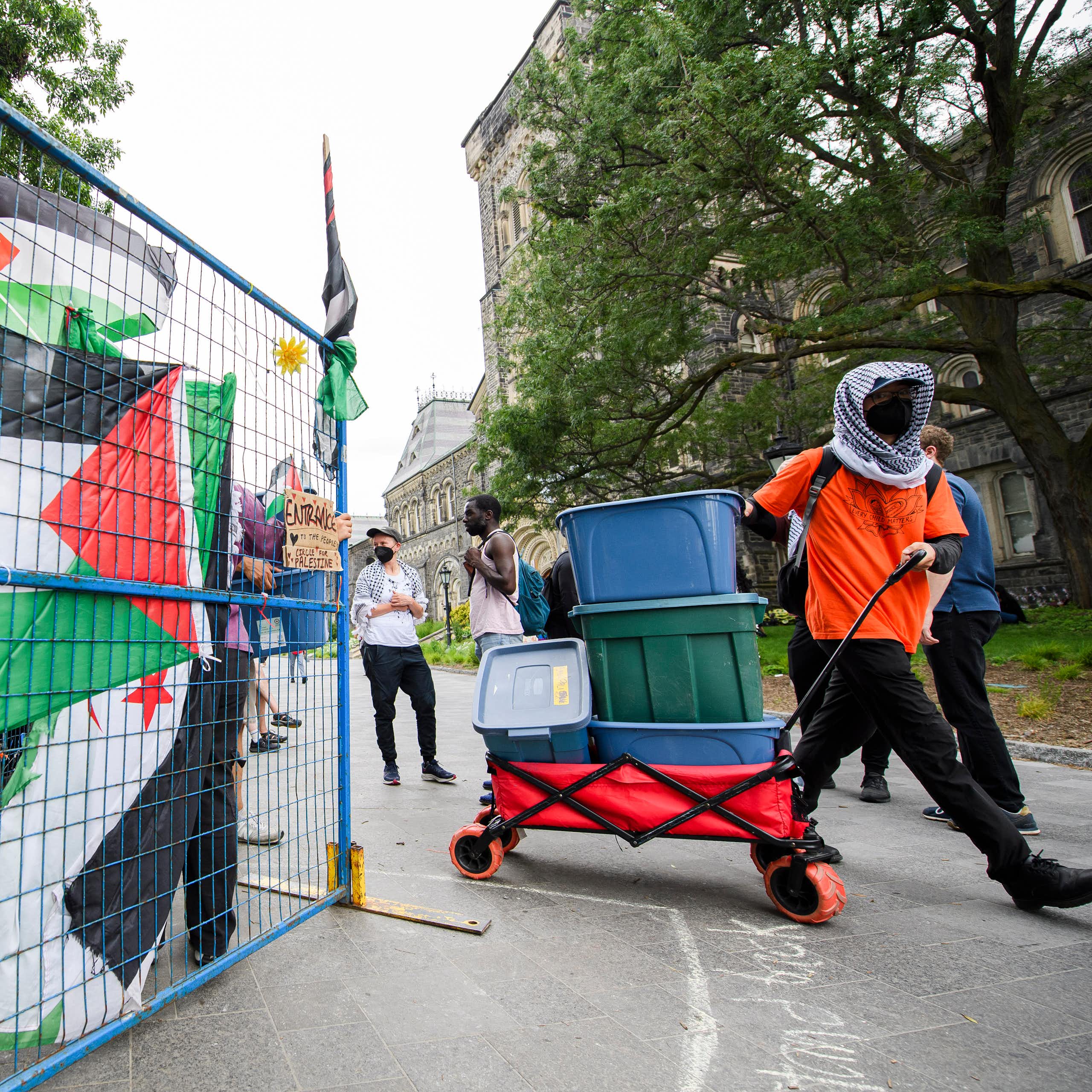 someone in an orange keffiyeh whose face is covered pulls a wagon of boxes past a Palestinian flag