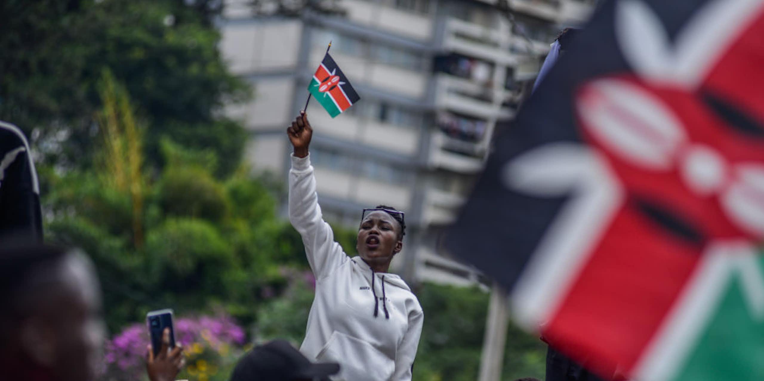 A woman in a grey jumper holding up a small flag with black, white, red and green stripes while facing a crowd of people who are also holding flags