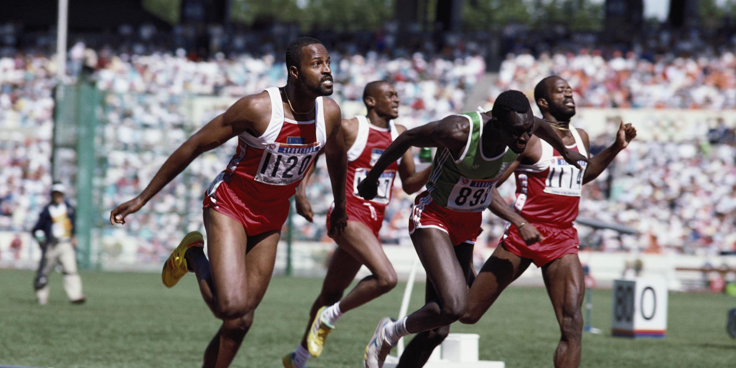 A vintage photo in colour of four men in athletics shorts and vests finishing a race on a track, some grimacing.