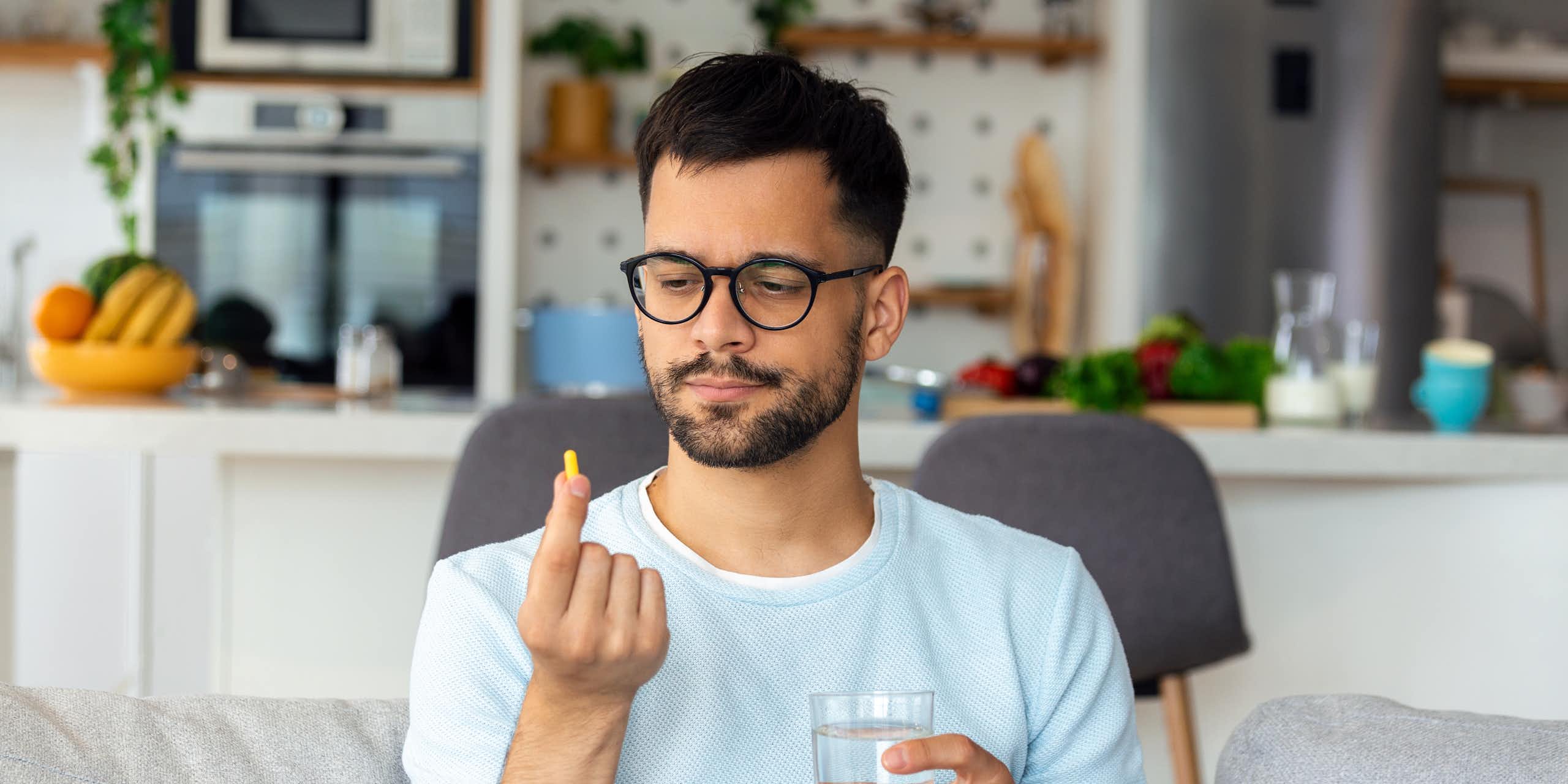 A man wearing glasses sitting on a sofa with a glass of water in one hand, looking at a pill in his other hand