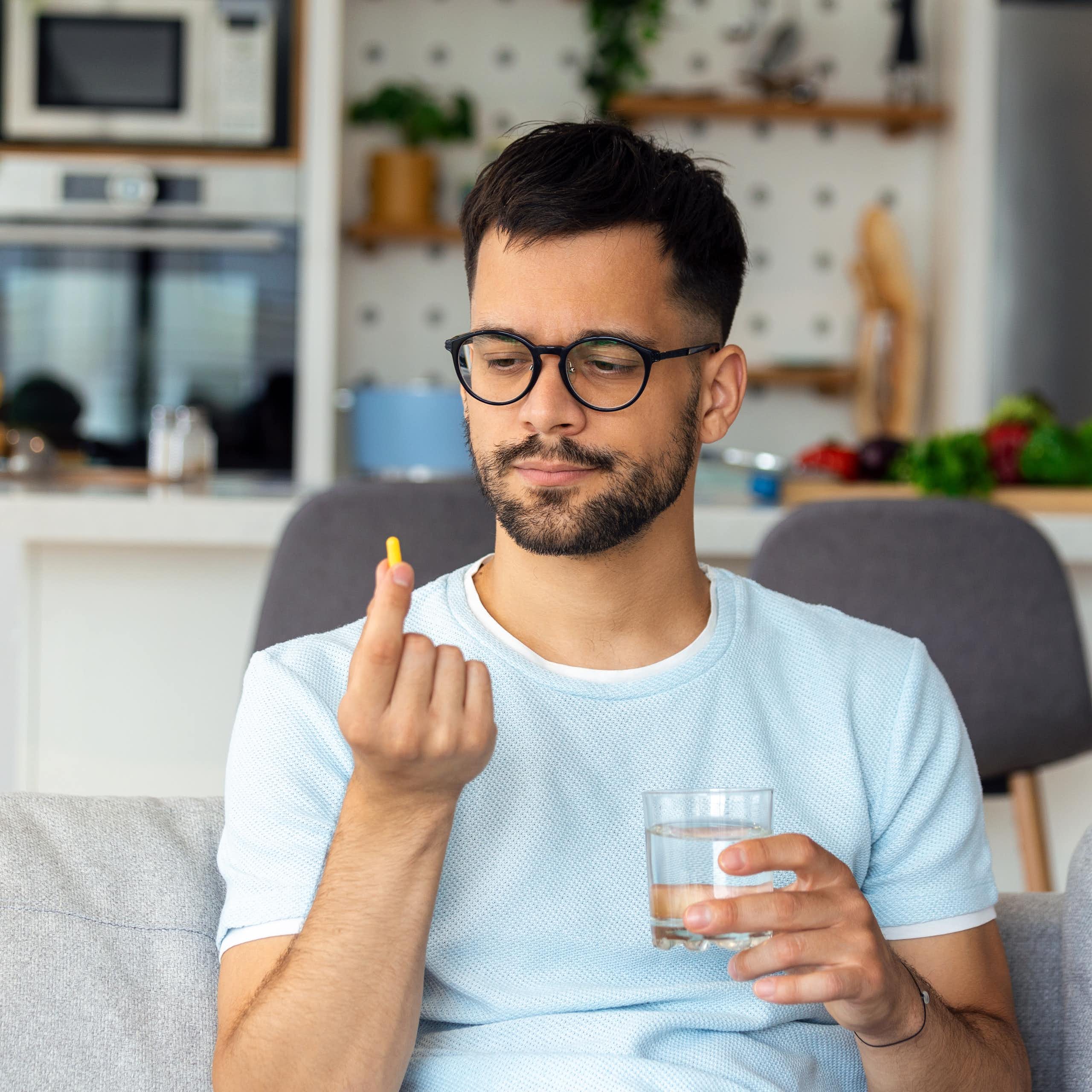 A man wearing glasses sitting on a sofa with a glass of water in one hand, looking at a pill in his other hand
