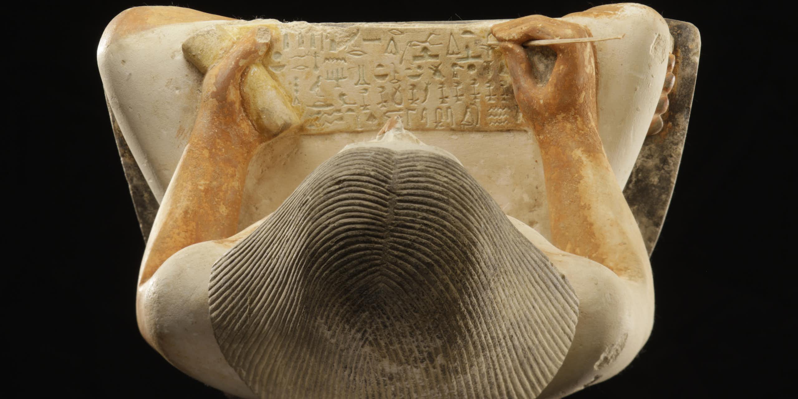 An overhead view of a statue of a person with a papyrus on their lap, engraving it with hieroglyphics