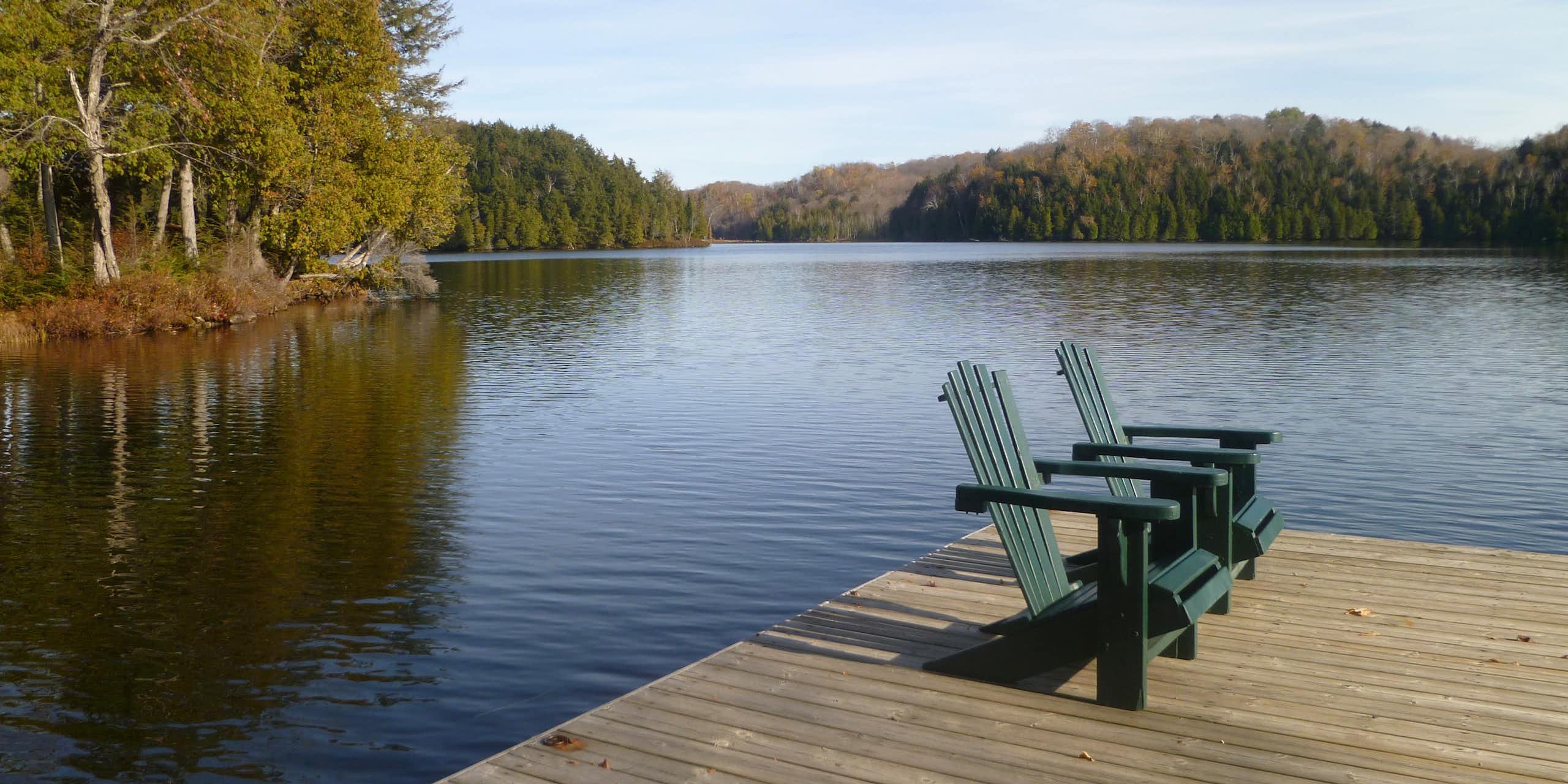 Two chairs stand side by side on the dock of a lake surrounded by a forest