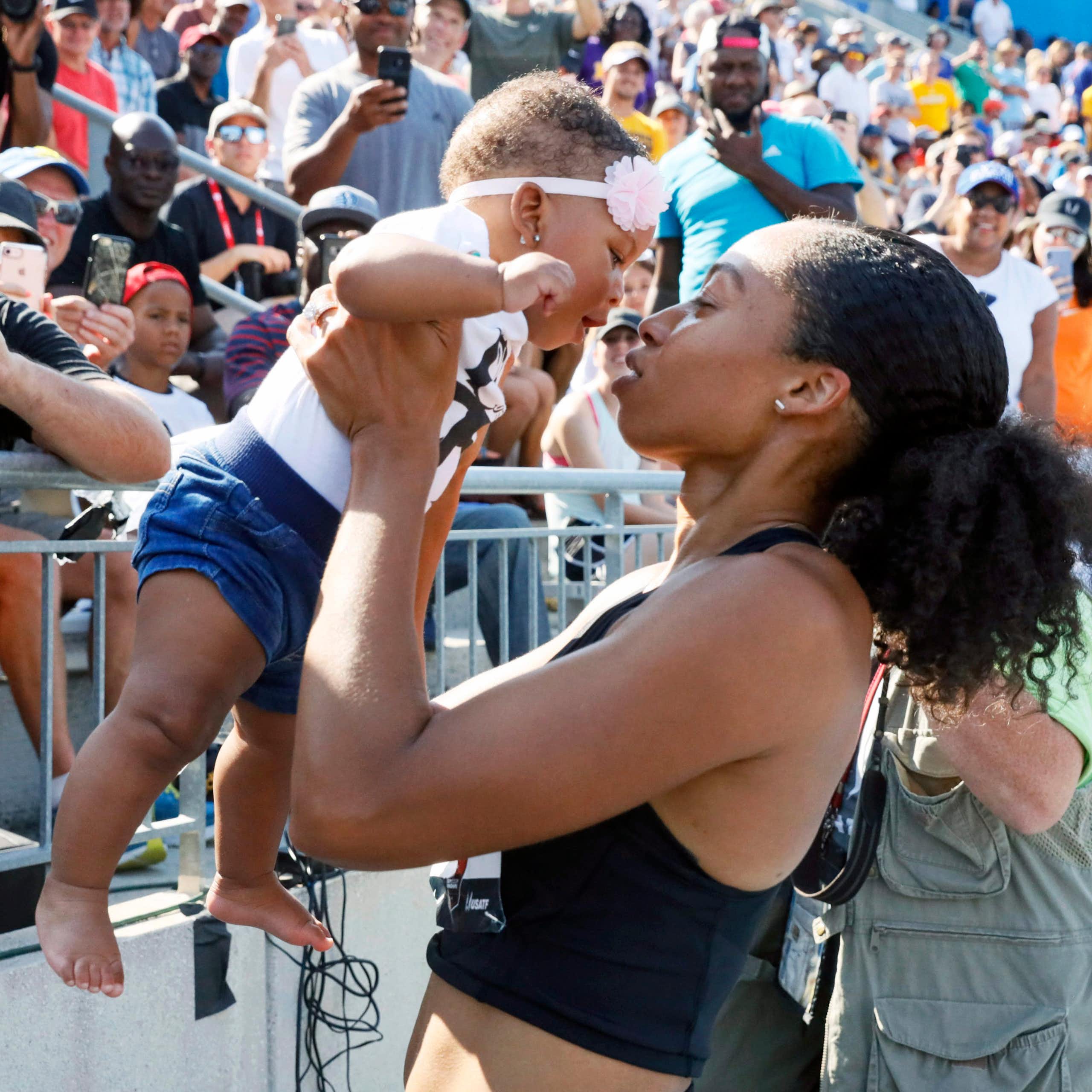 A Black woman in a cropped tank top lifts a baby up, who is wearing a headband with a flower on it, while standing in front of a set of bleachers full of people
