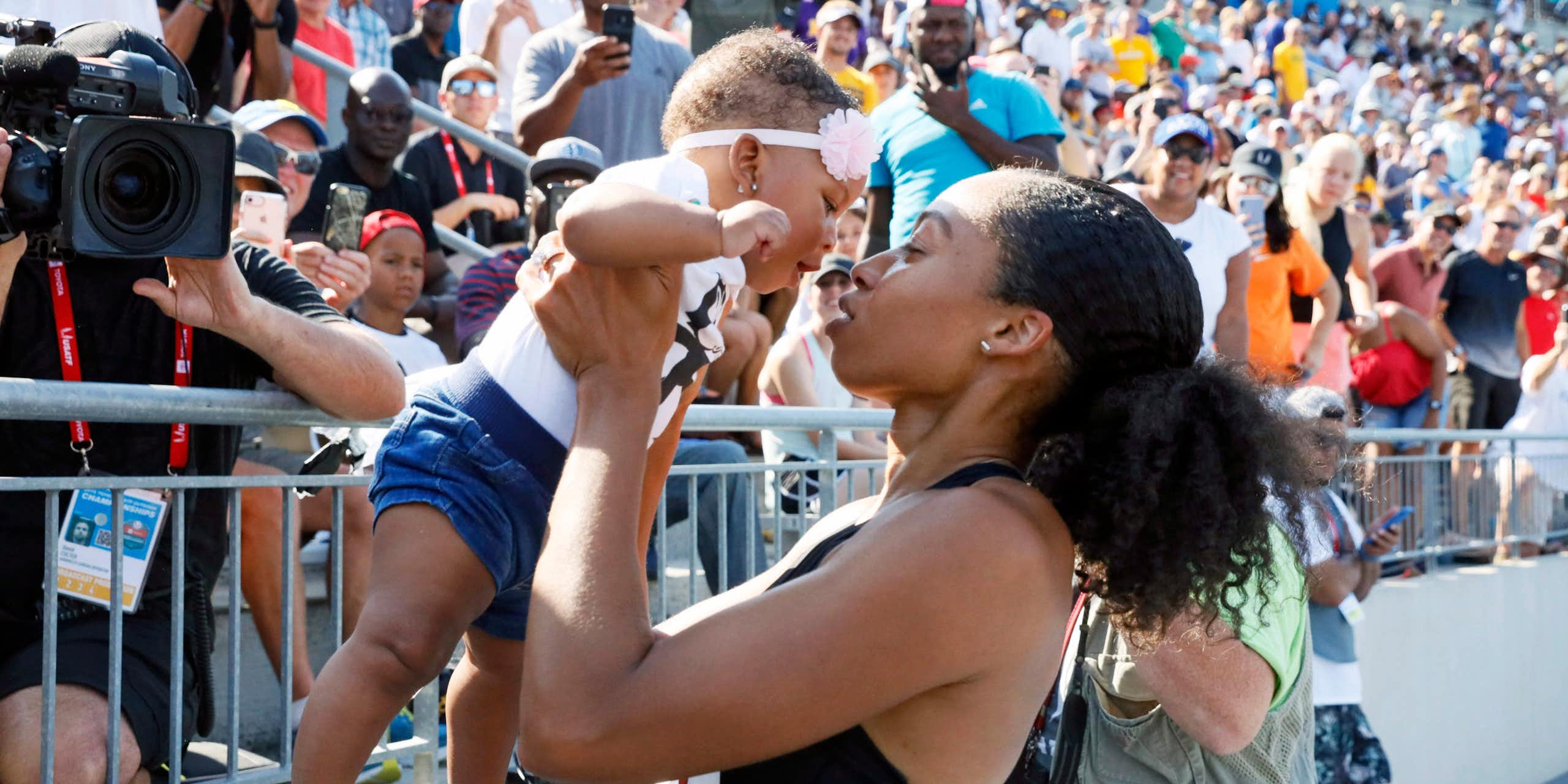 A Black woman in a cropped tank top lifts a baby up, who is wearing a headband with a flower on it, while standing in front of a set of bleachers full of people
