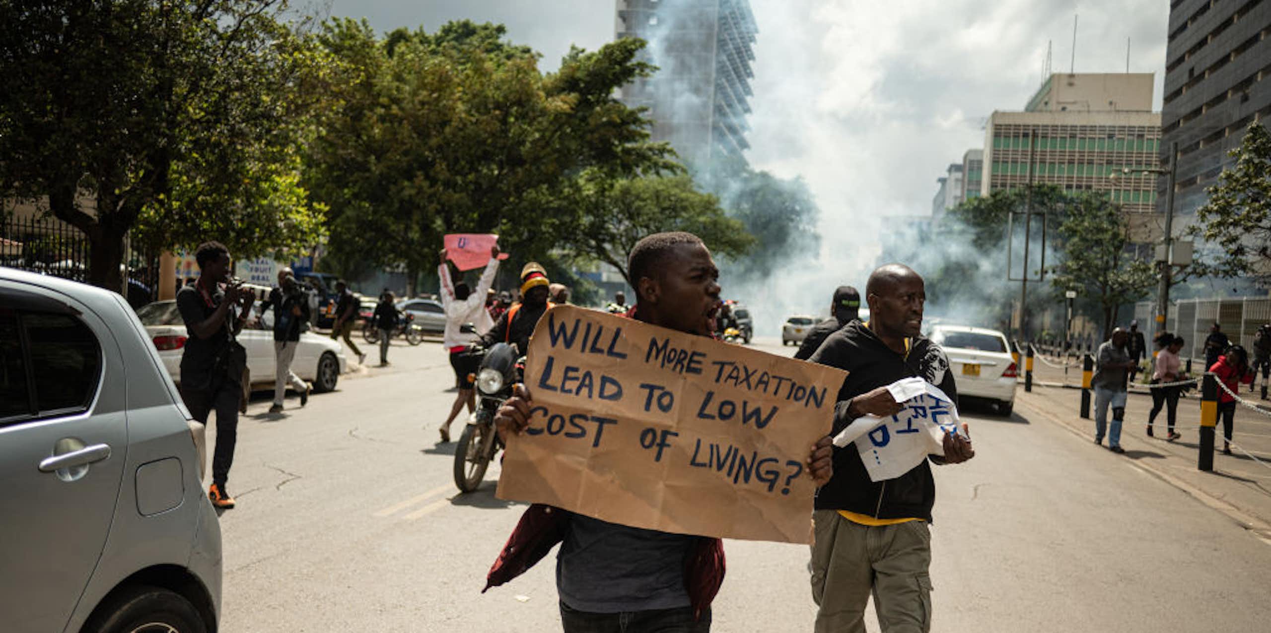 Protesters waving placards on a city street with a cloud of tear gas rising behind them.