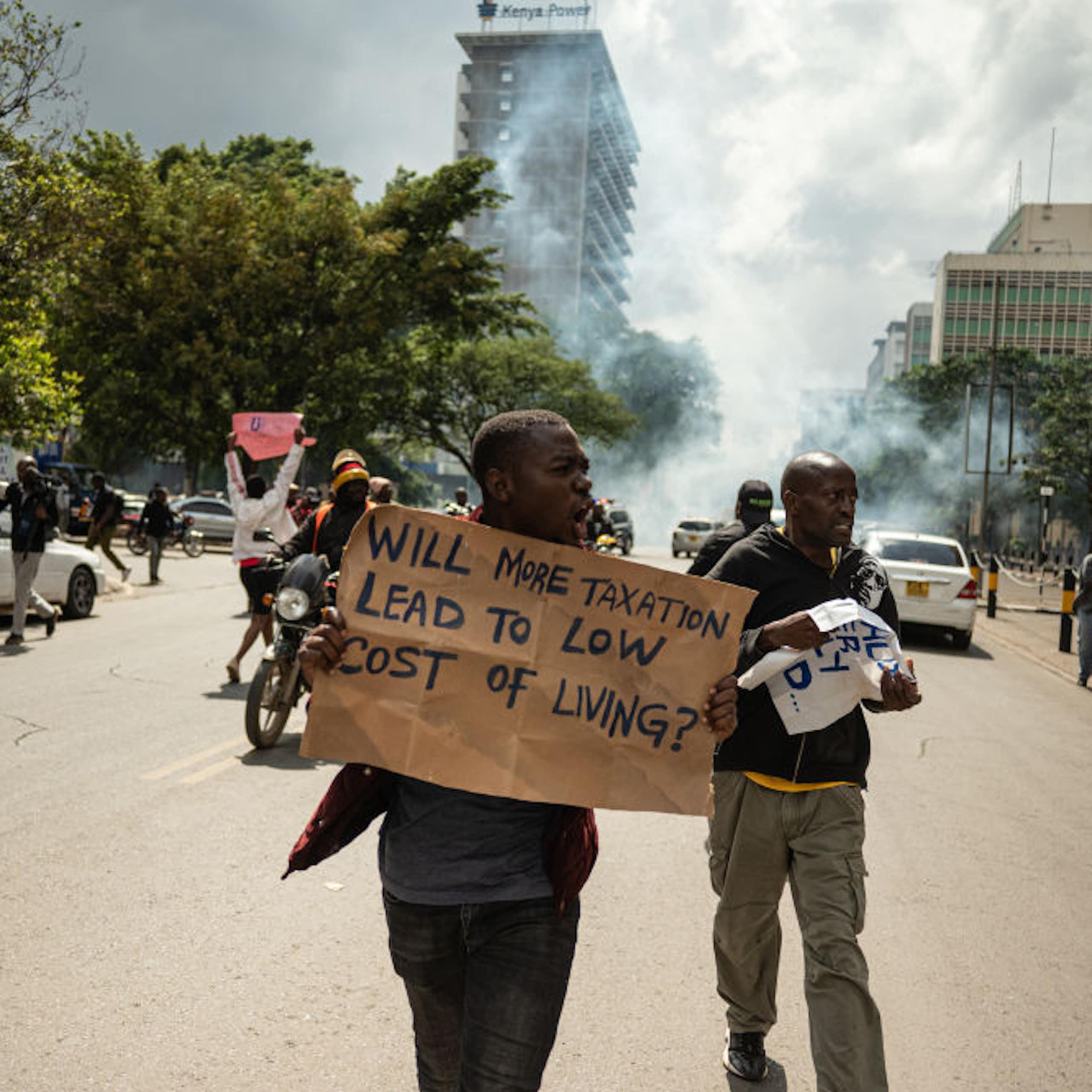Protesters waving placards on a city street with a cloud of tear gas rising behind them.