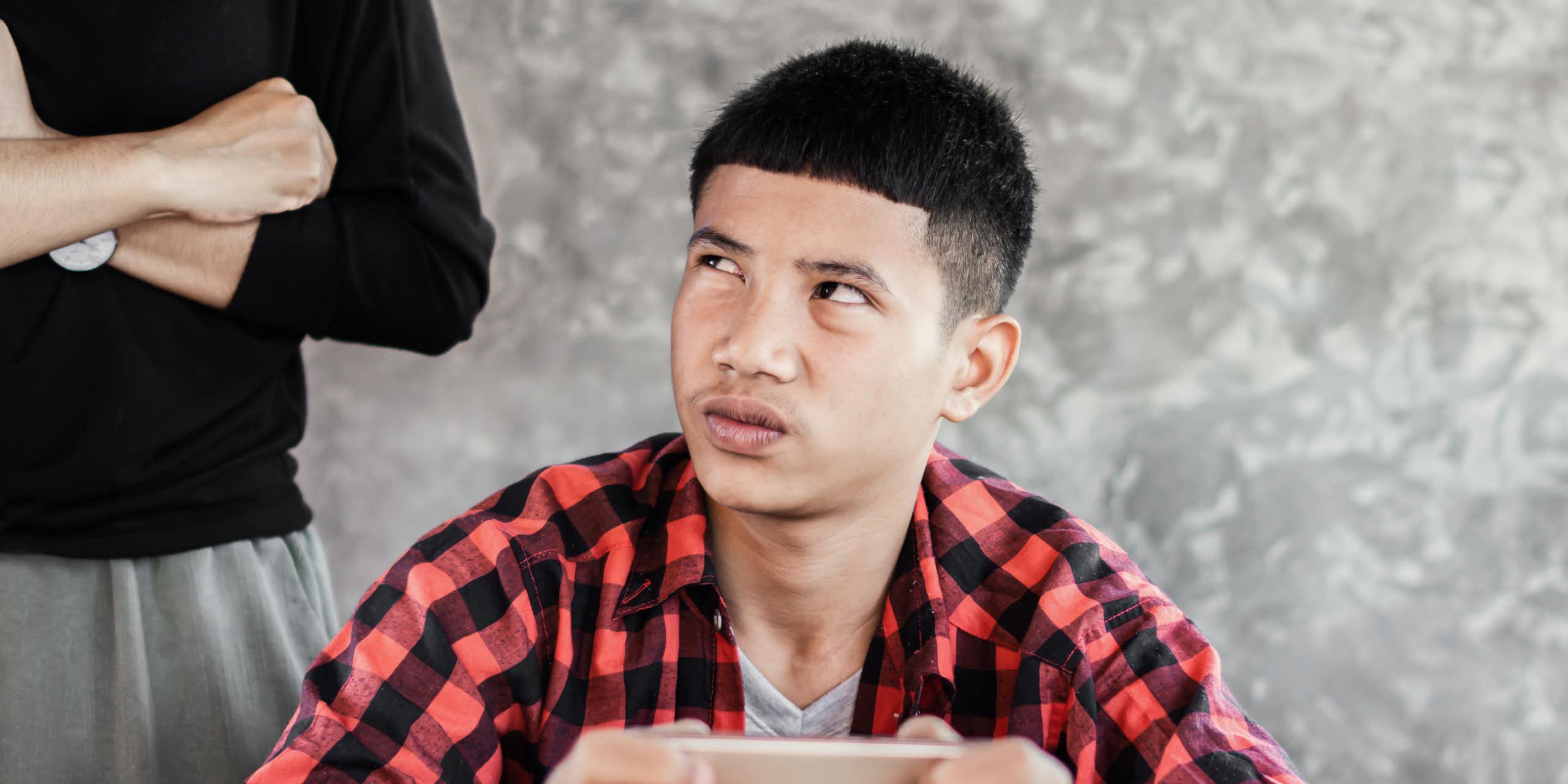 a boy holds an iphone and looks up at an adult standing behind him with their arms crossed