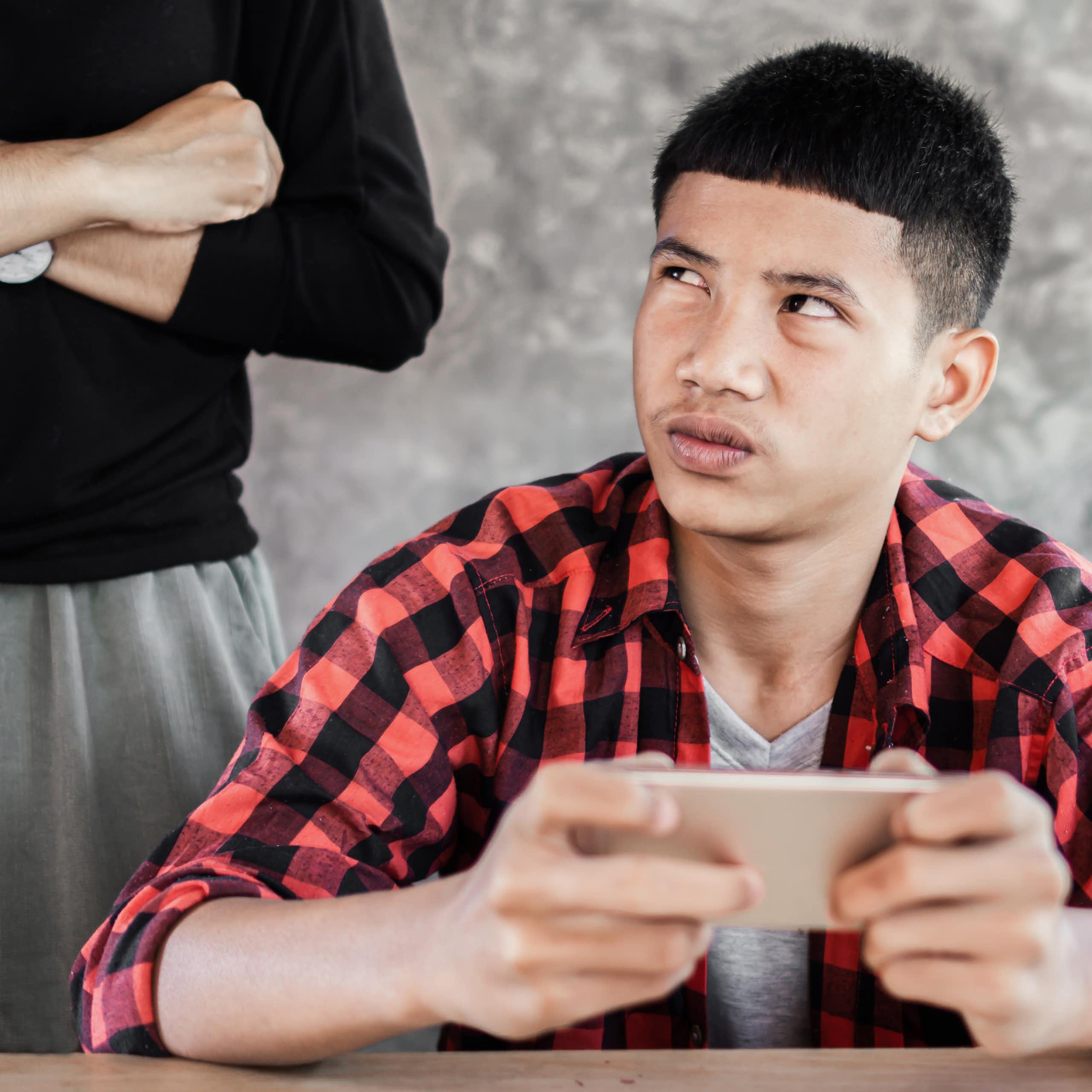 a boy holds an iphone and looks up at an adult standing behind him with their arms crossed