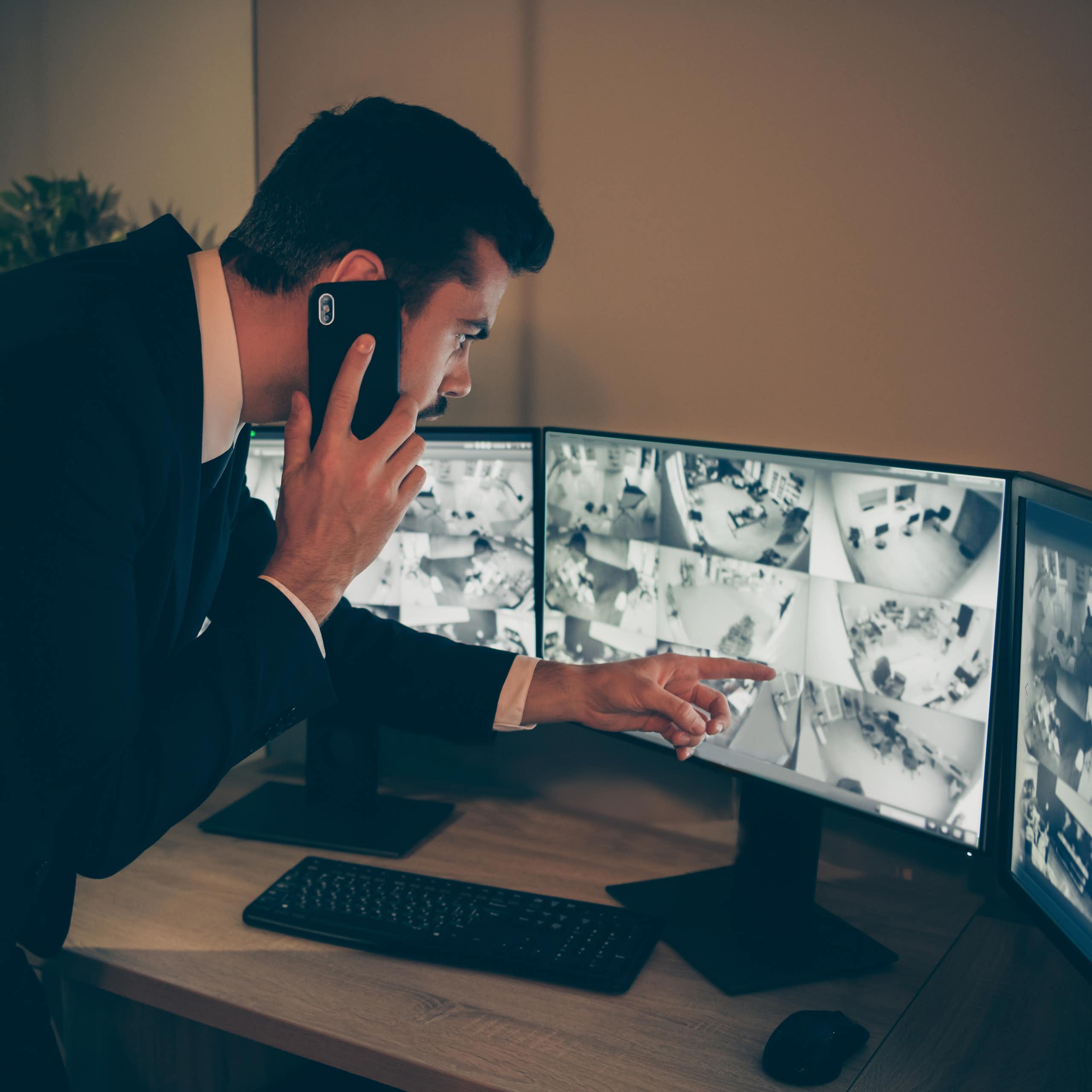 A man points at a CCTV screen while talking on a cellphone.