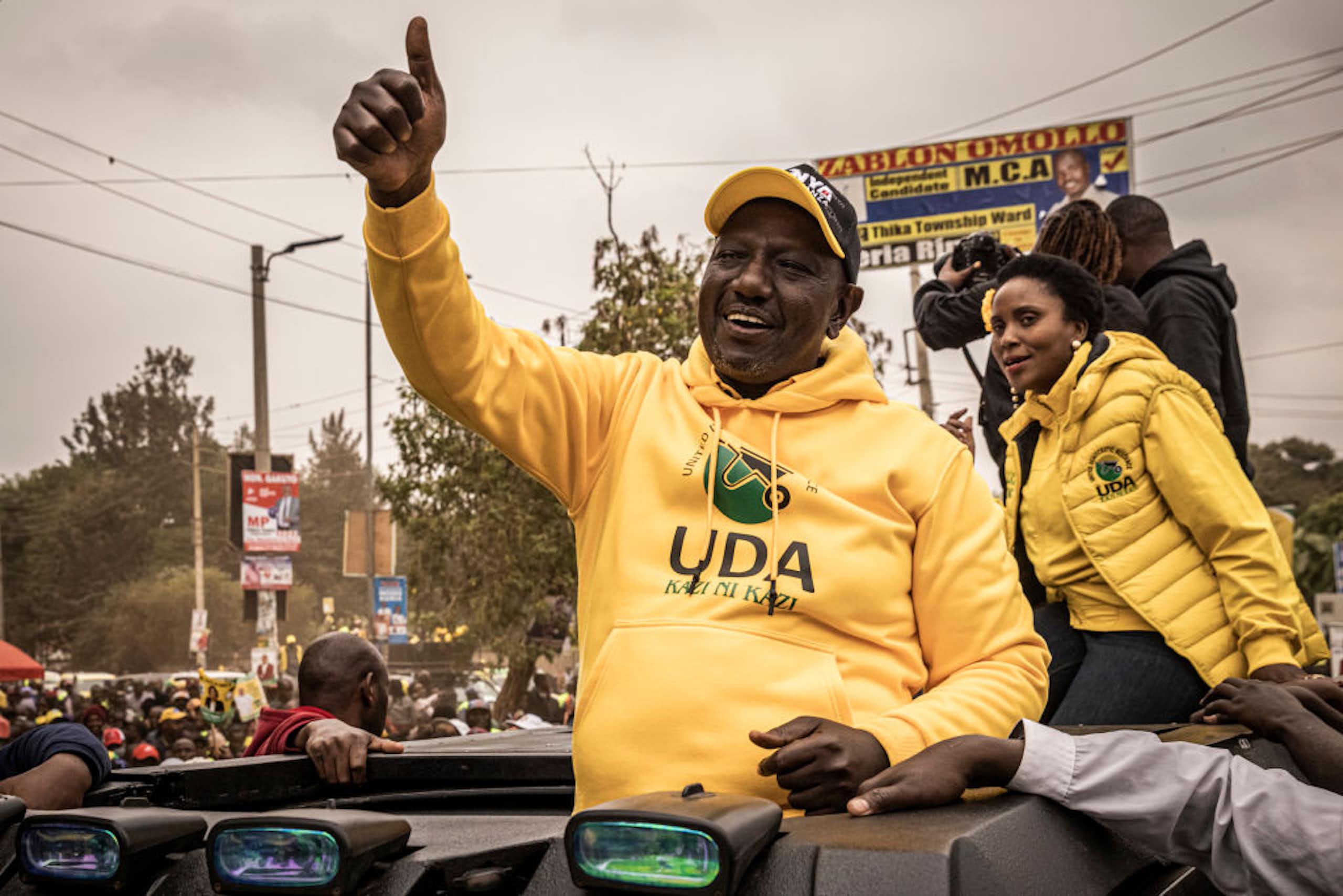 A man holding up through the roof of a car with one hand holding up a thumbs up with a crowd of people int the background 