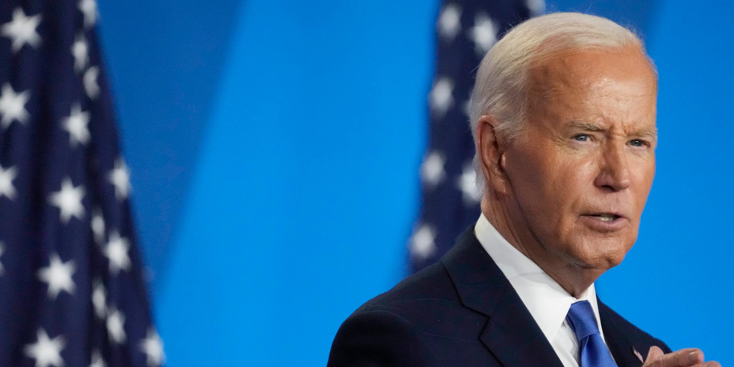 A white haired man in blue blazer standing in front of two flags.