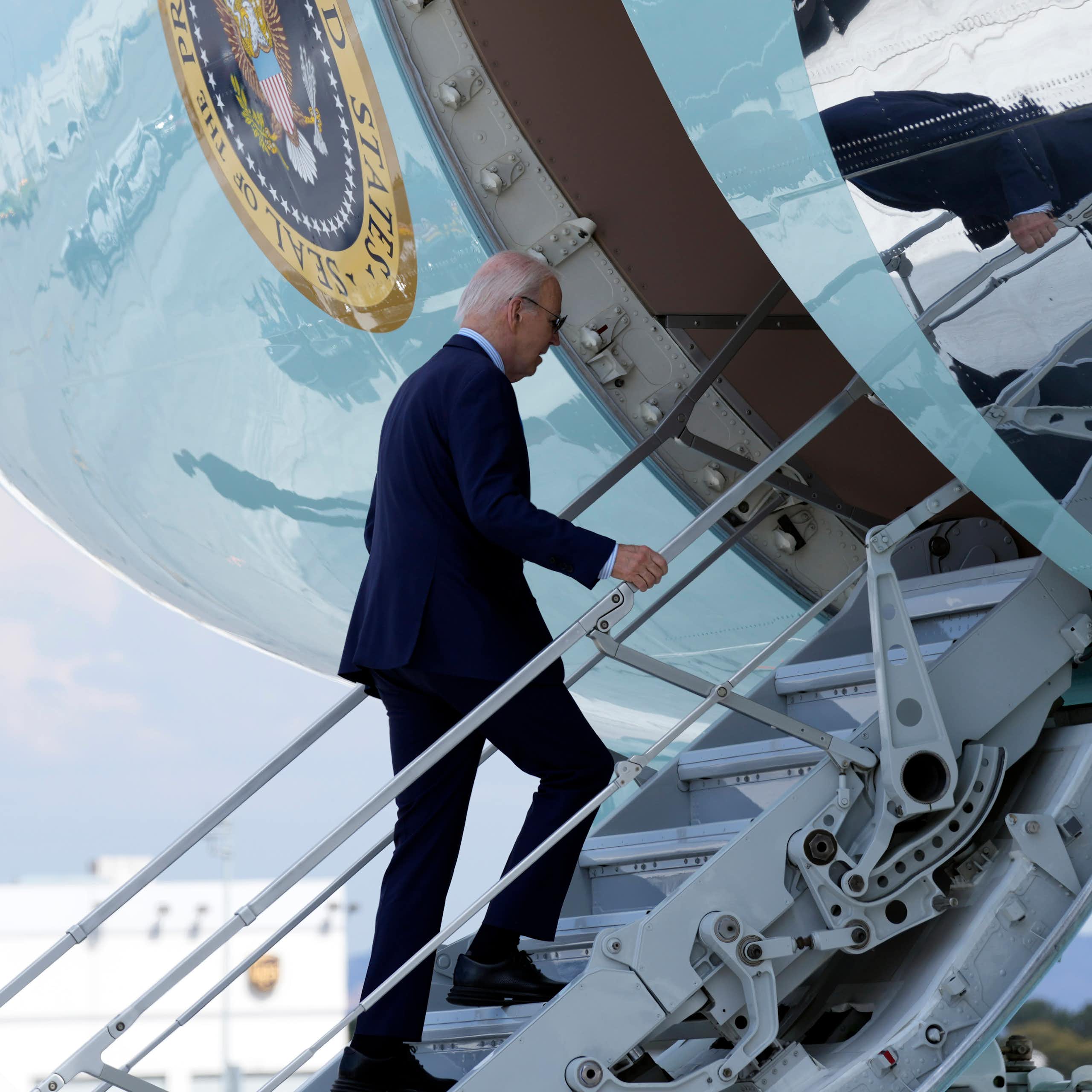 An aging man with thinning grey hair a blue suit walks up the steps to an airliner.