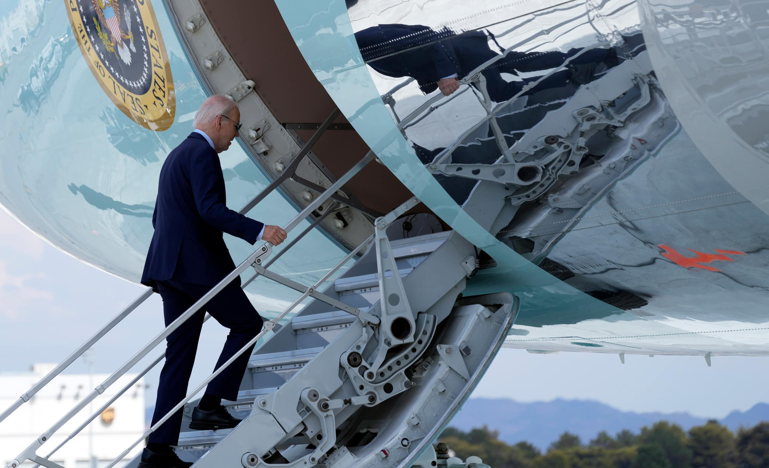 An aging man with thinning grey hair a blue suit walks up the steps to an airliner.