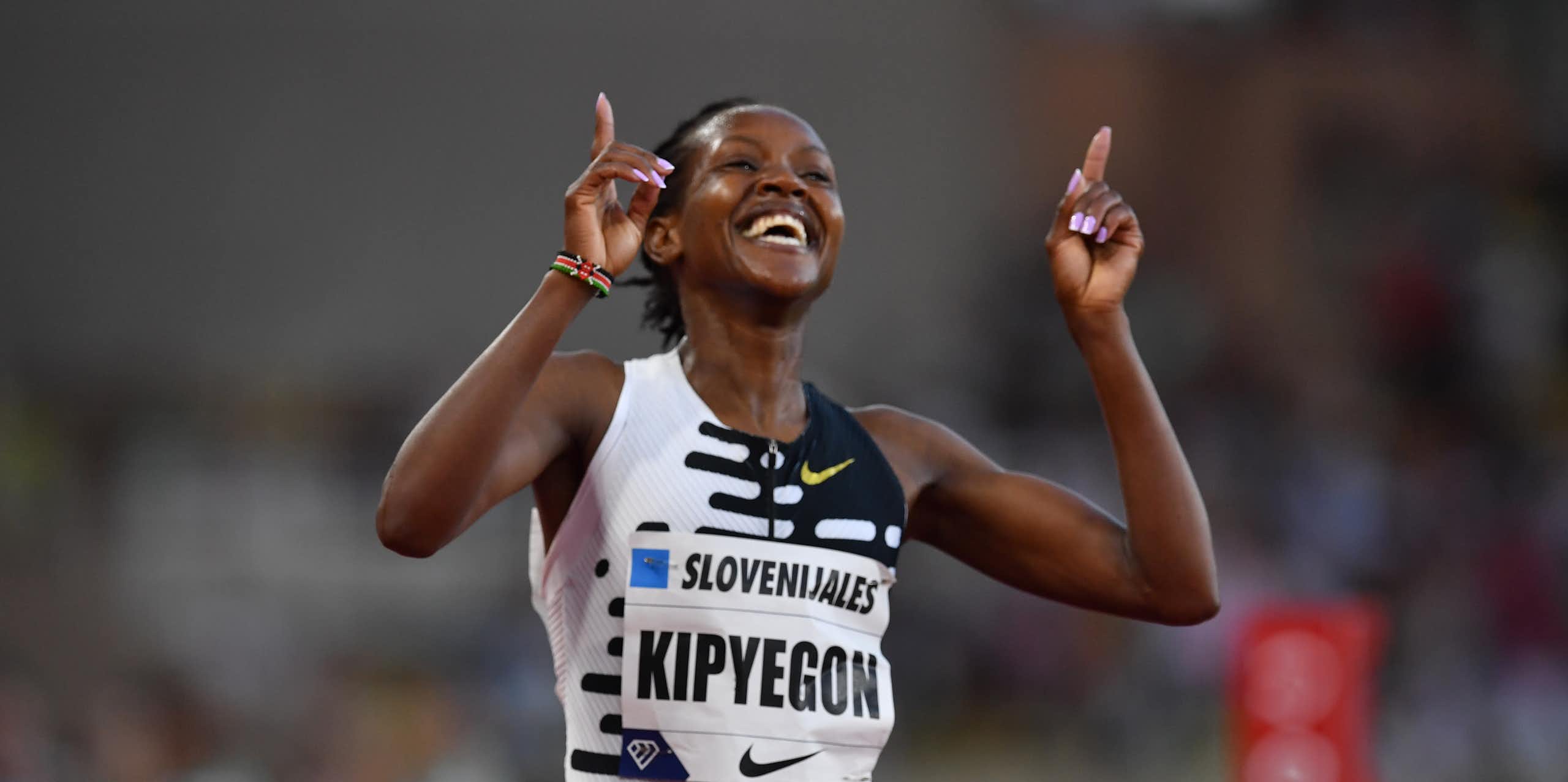 An African woman in athletics vest beams with raised arms, index fingers pointing upwards in victory.