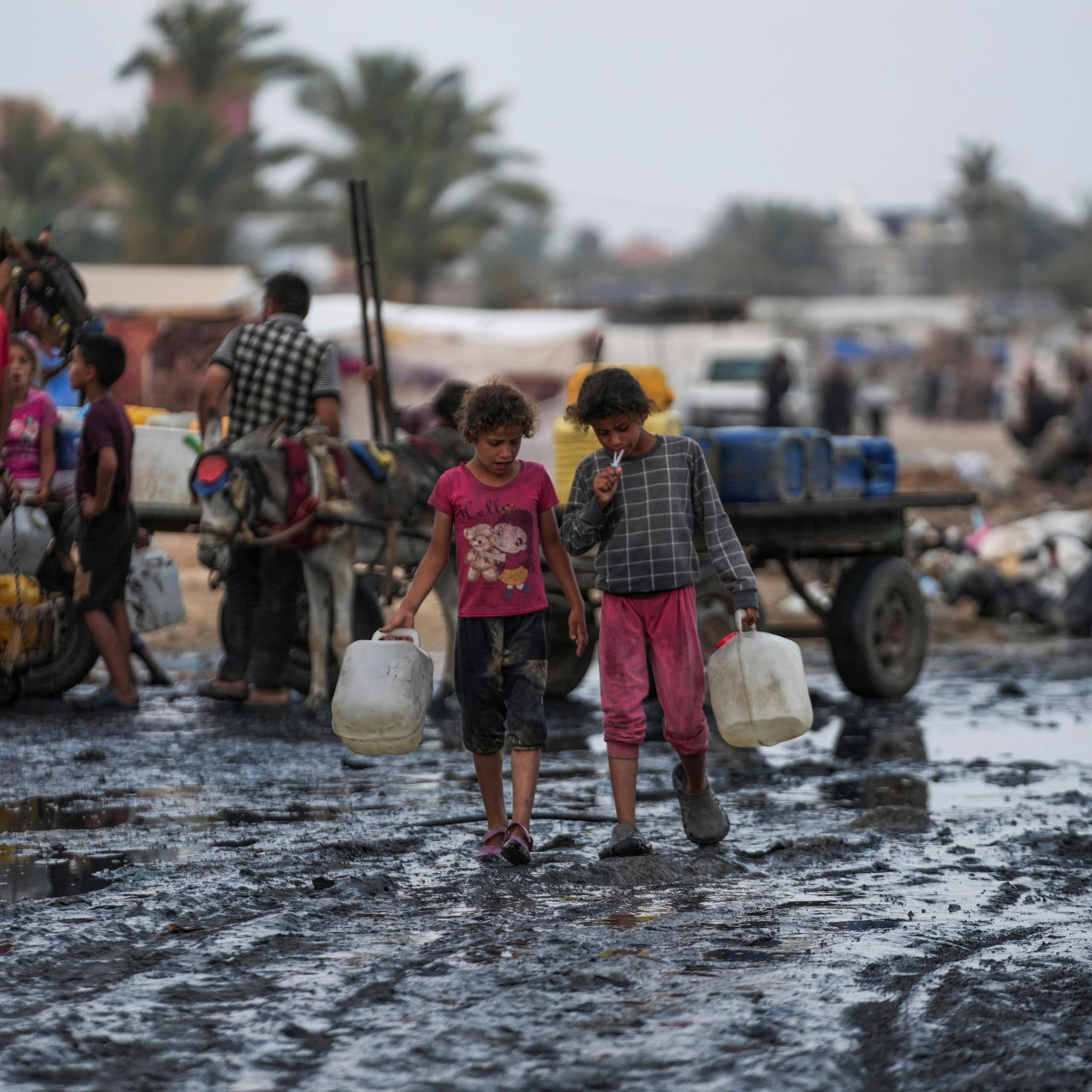 children carrying water bottles walk through muddy area