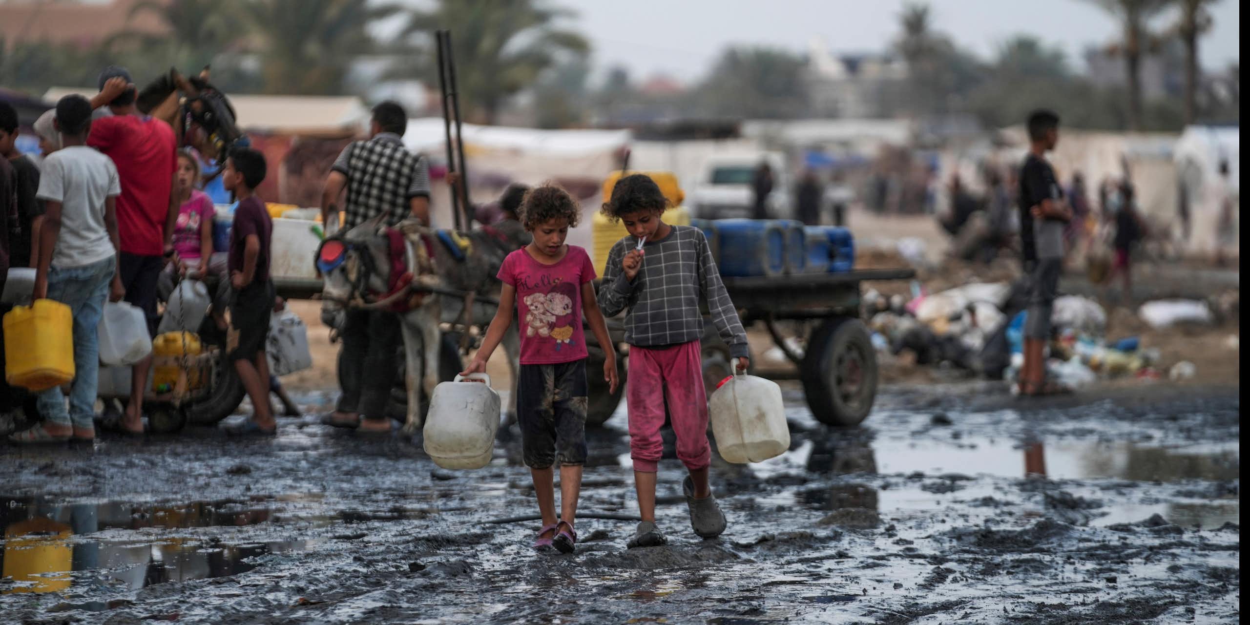 children carrying water bottles walk through muddy area