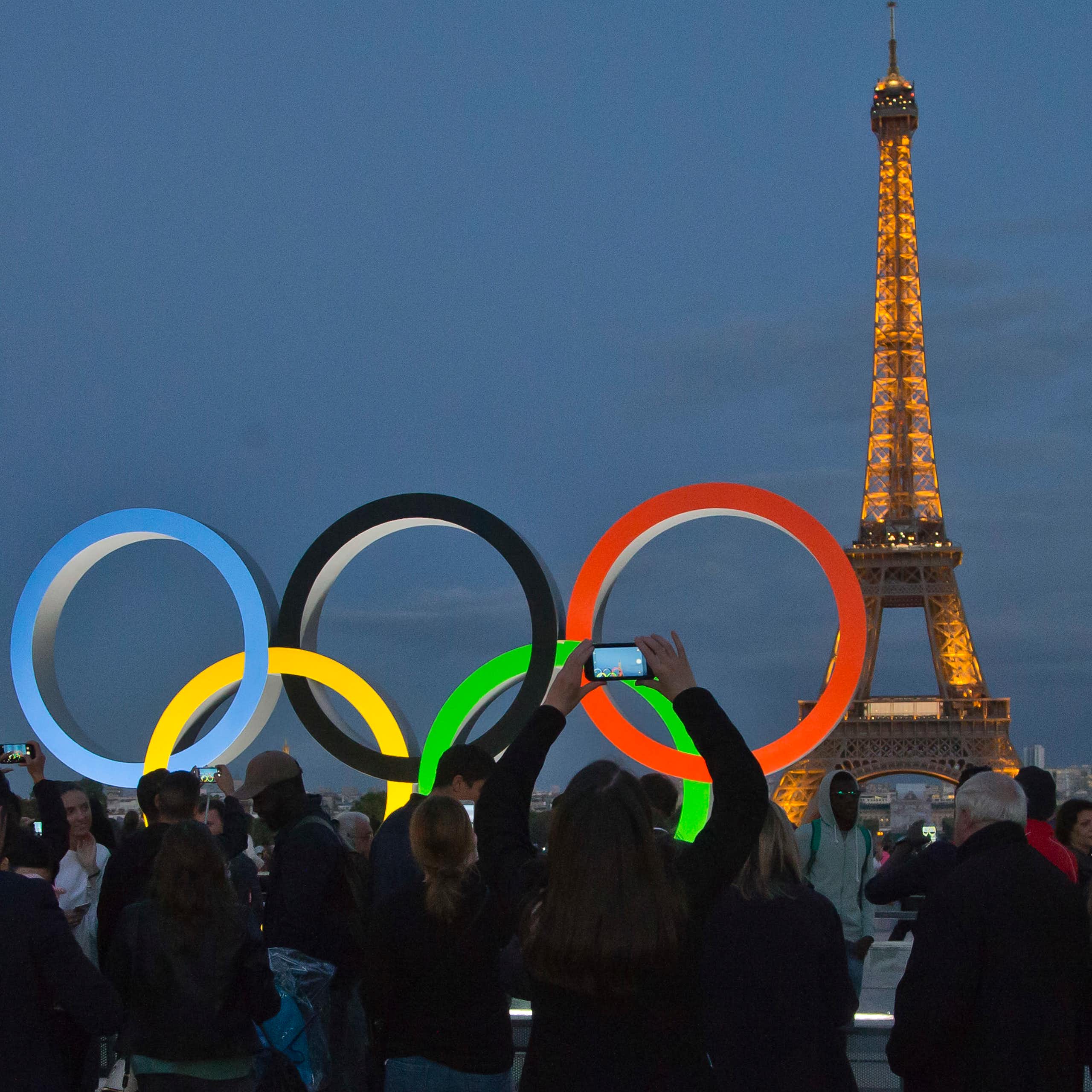 Os anéis olímpicos iluminados com a Torre Eiffel ao fundo. Pessoas em silhueta estão tirando fotos.
