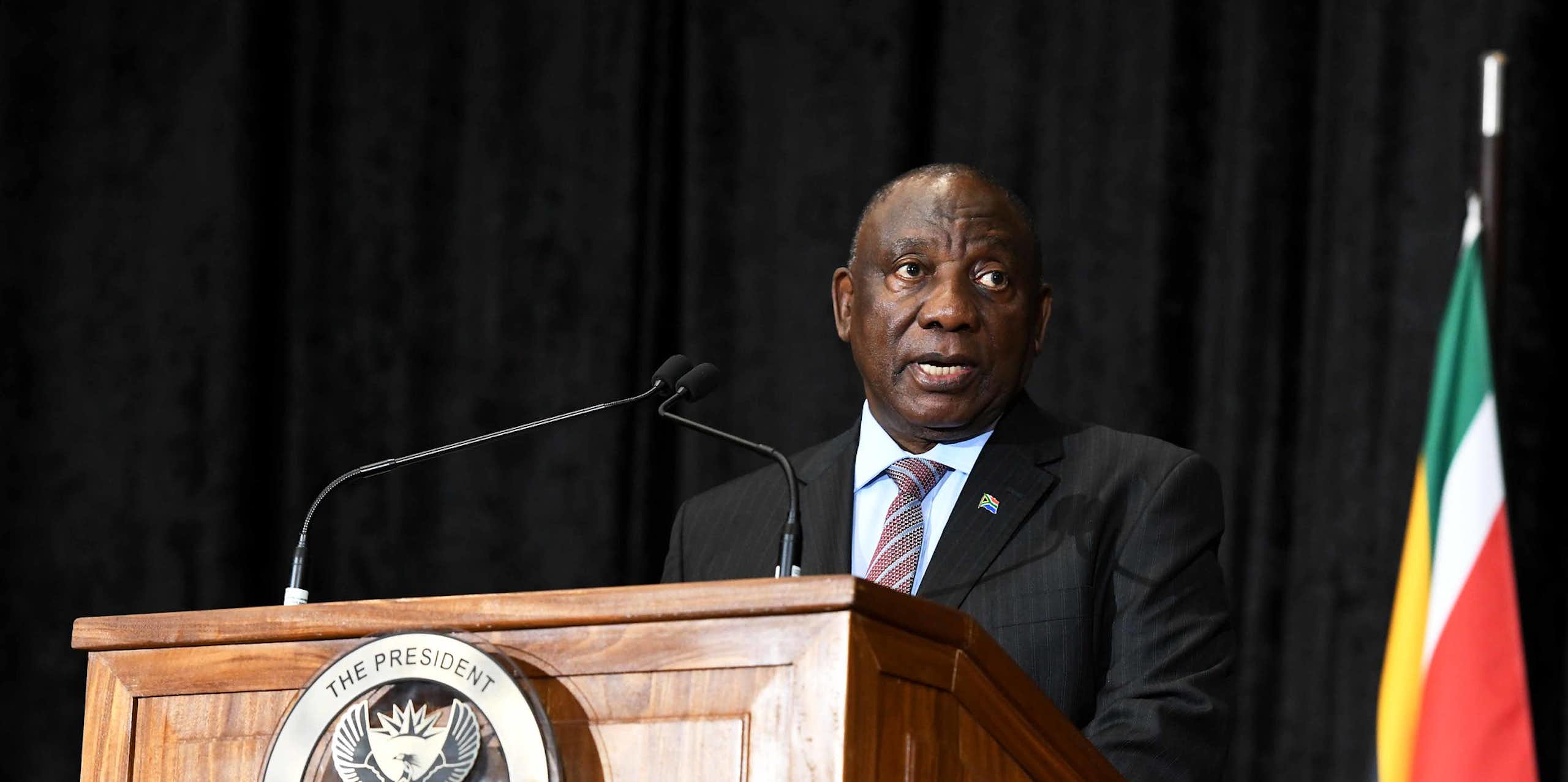 A man wearing a jacket and tie speaks at a podium bearing the South African coat of arms, next to the South African flag on his left.