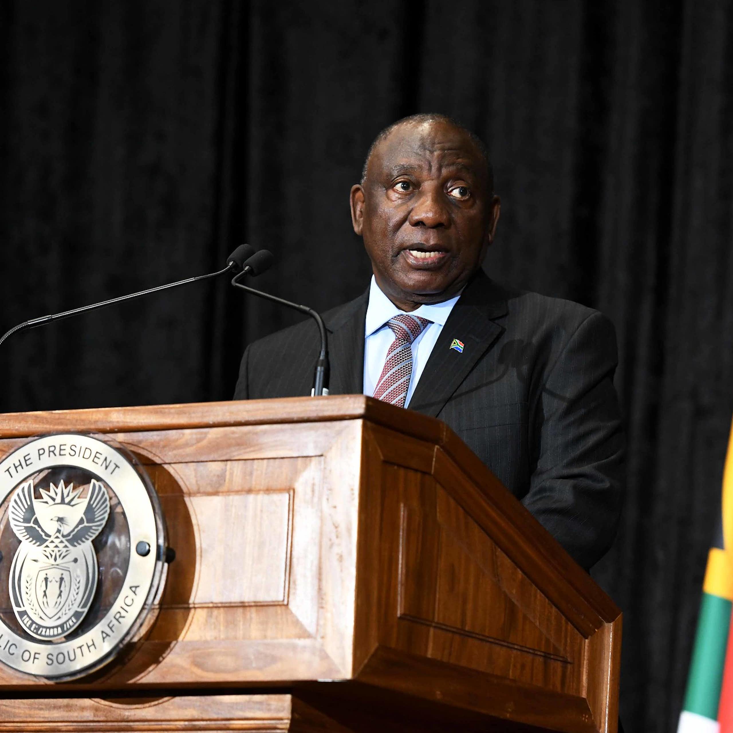 A man wearing a jacket and tie speaks at a podium bearing the South African coat of arms, next to the South African flag on his left.