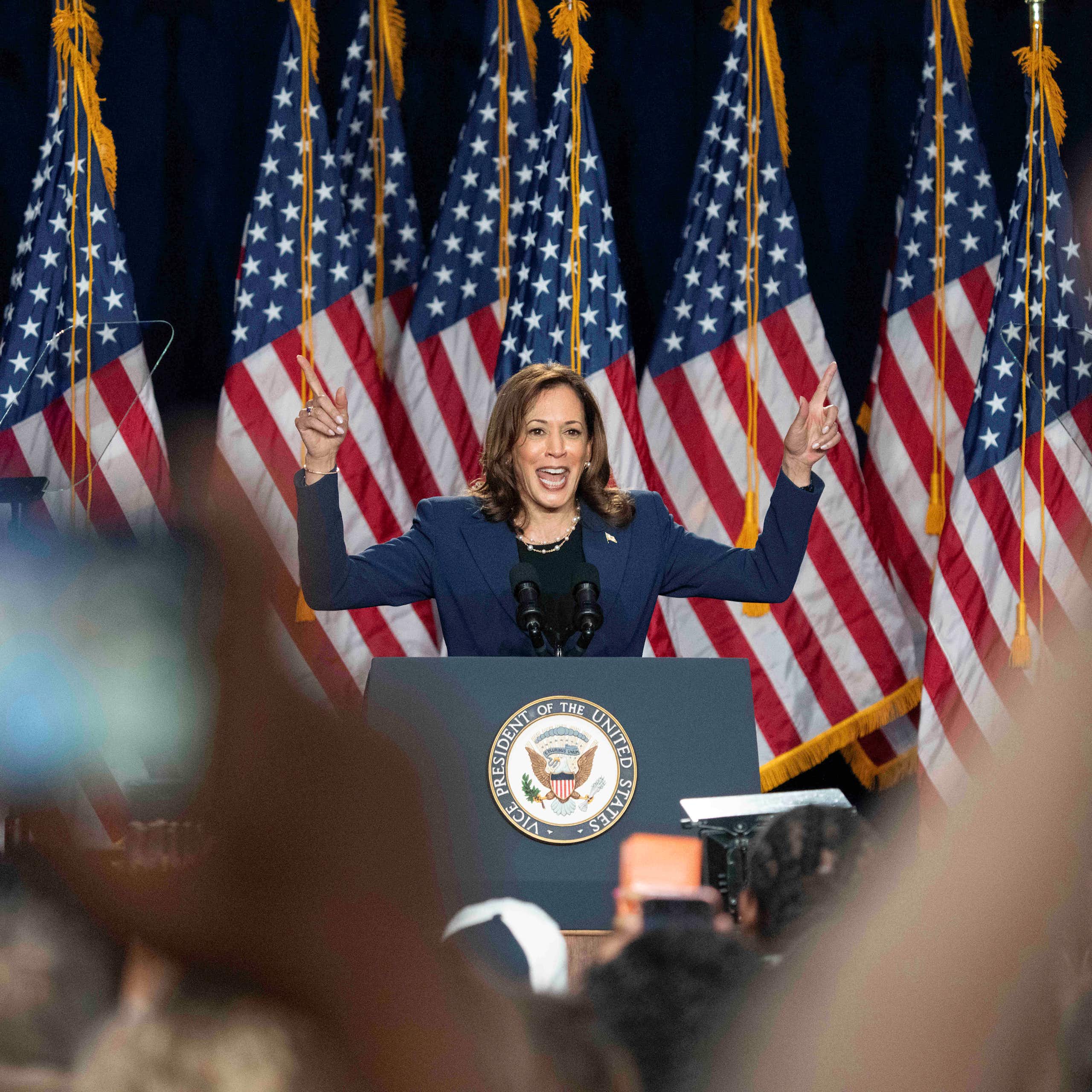 A smiling dark-haired woman raises her hands while speaking in front of a crowd, a row of American flags behind her.
