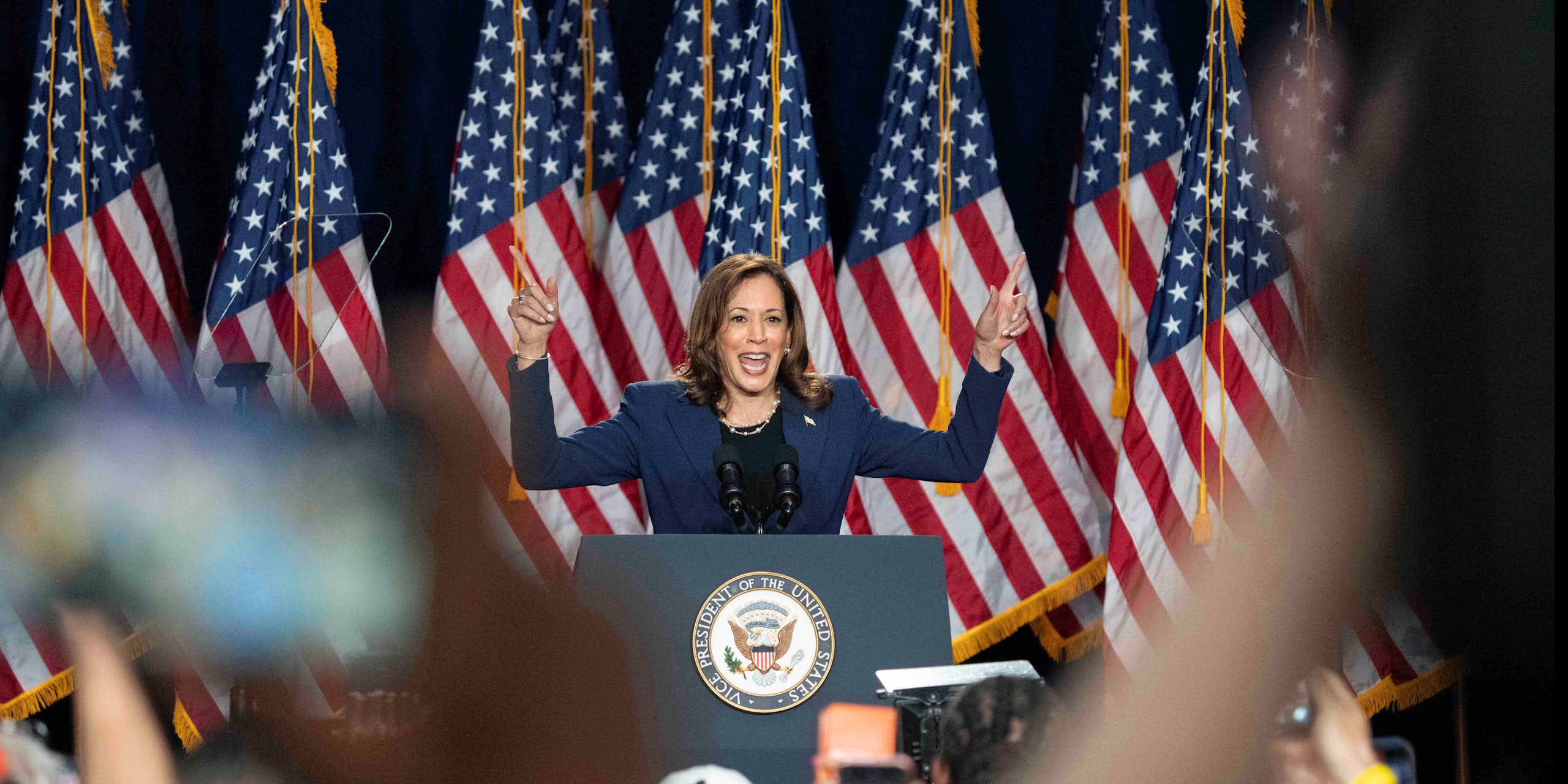 A smiling dark-haired woman raises her hands while speaking in front of a crowd, a row of American flags behind her.