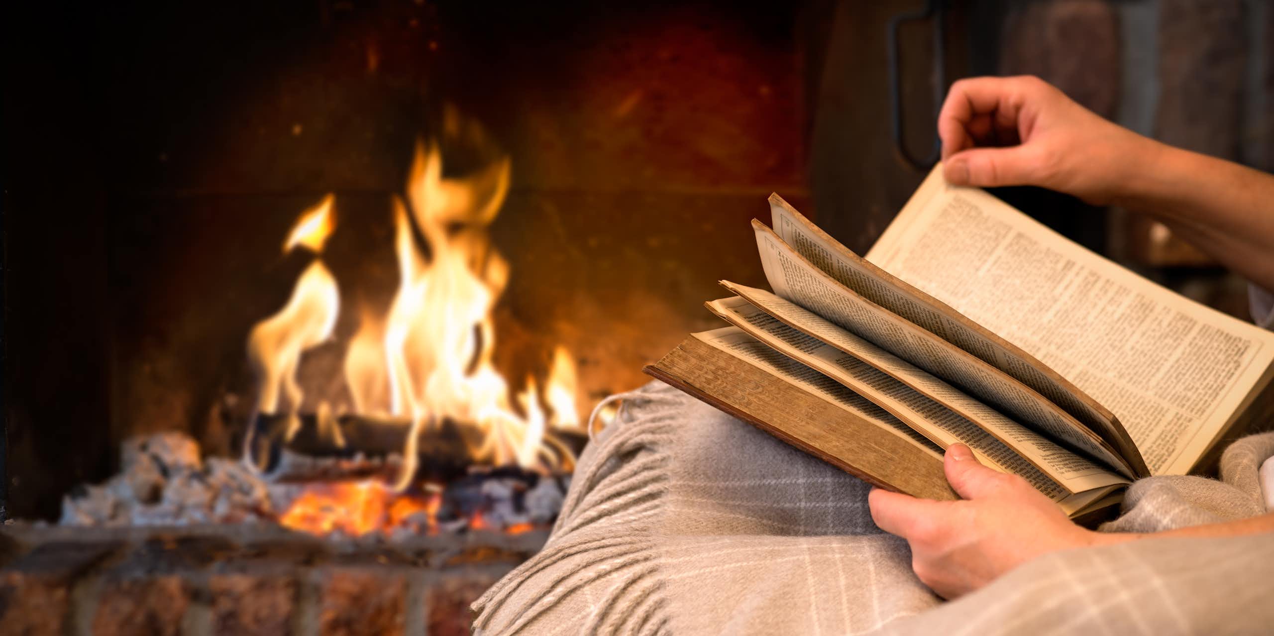 A person reads a book in front of an open fireplace