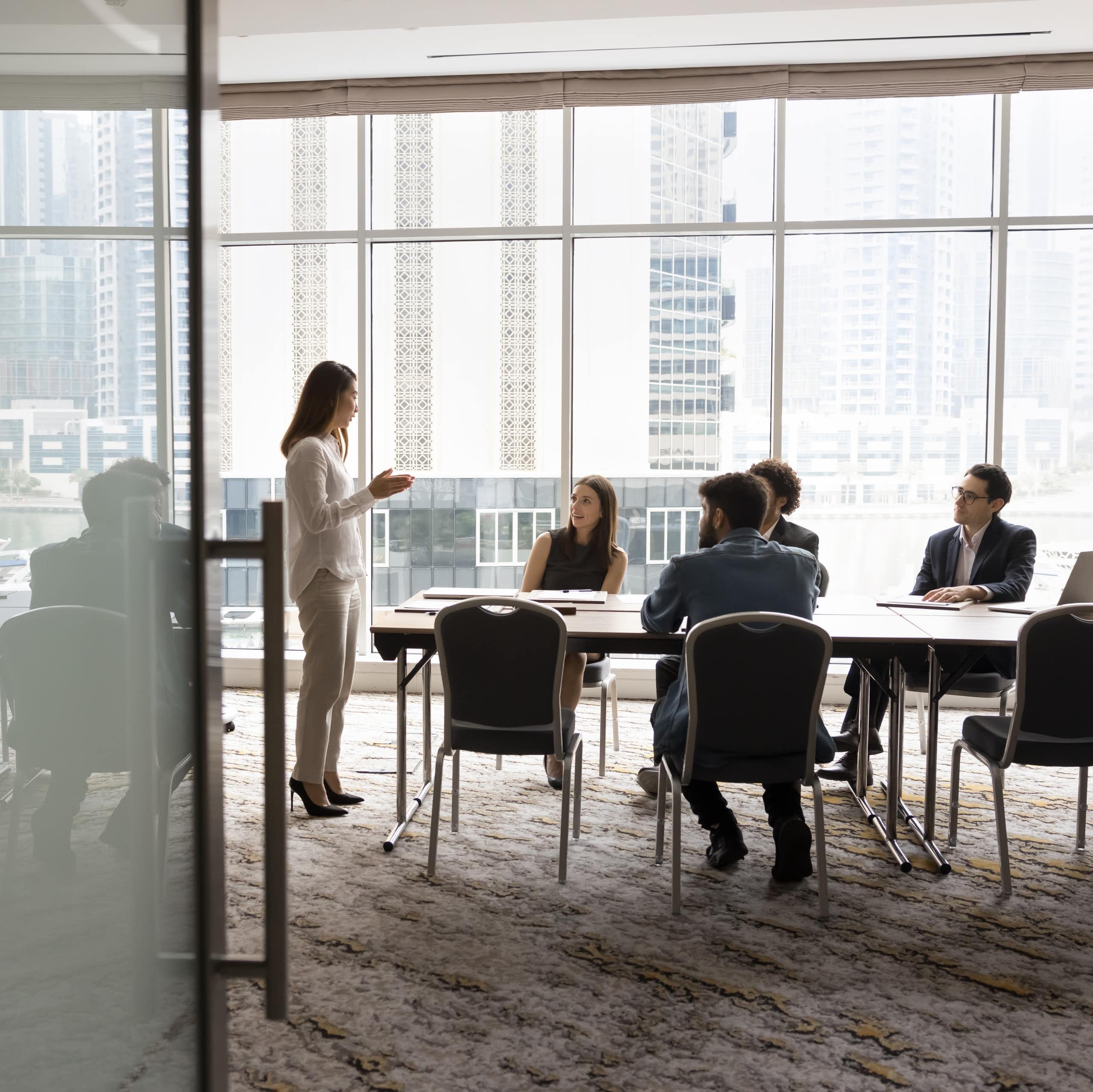 Wide shot of a group of people sitting at a conference table, looking up at a woman who is speaking to them