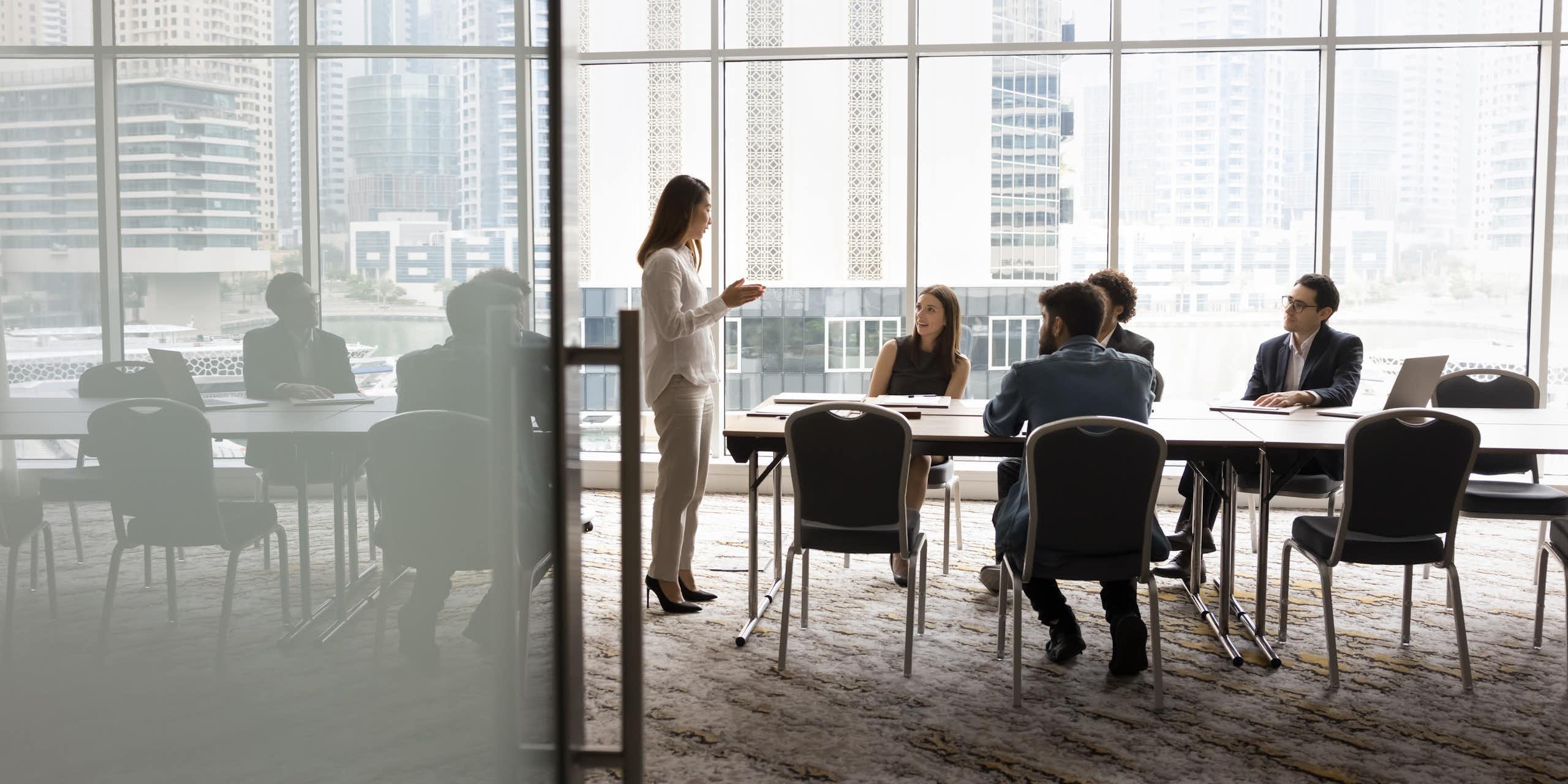 Wide shot of a group of people sitting at a conference table, looking up at a woman who is speaking to them