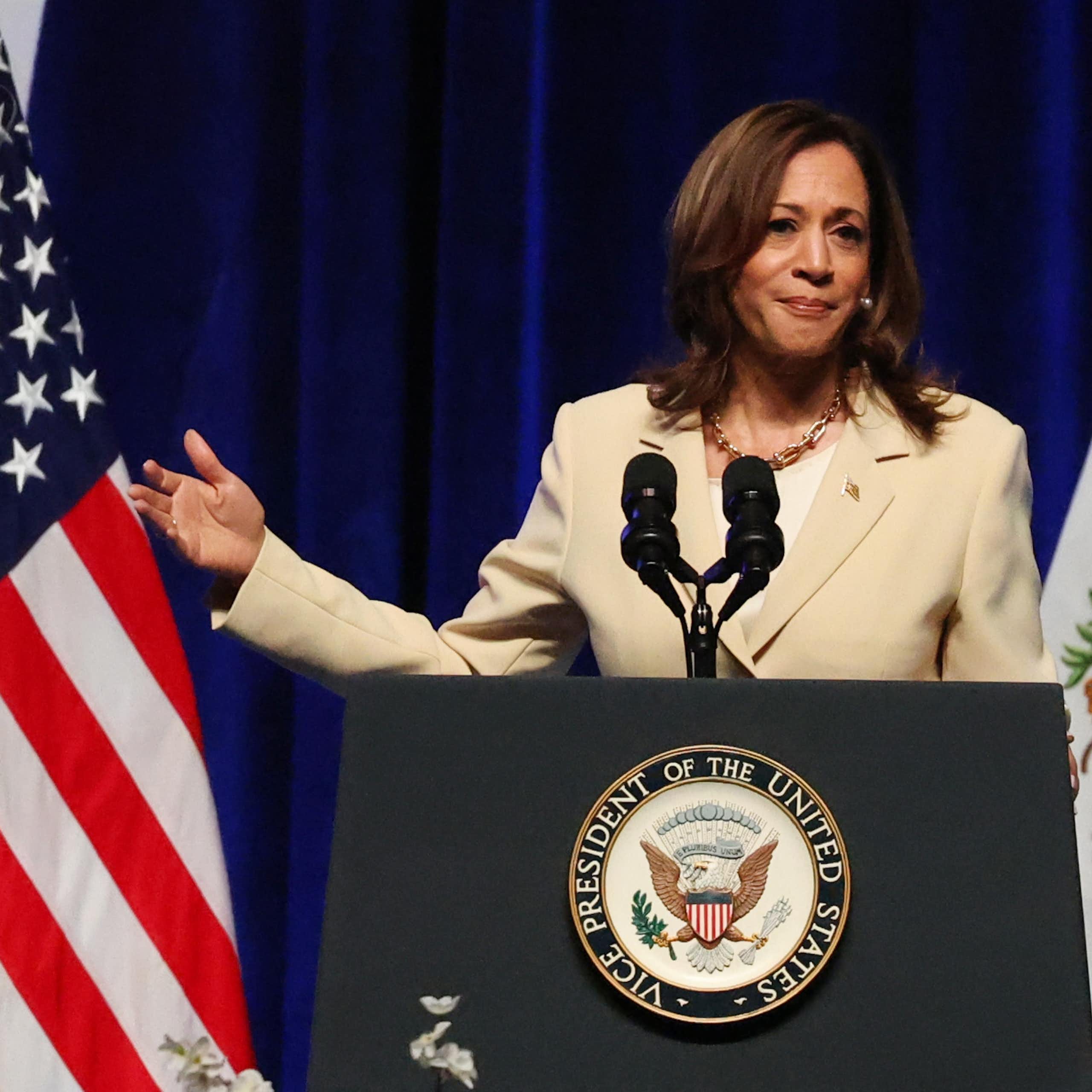 US vice-president Kamala Harris stadns at a podium gesturing towards an American flag.