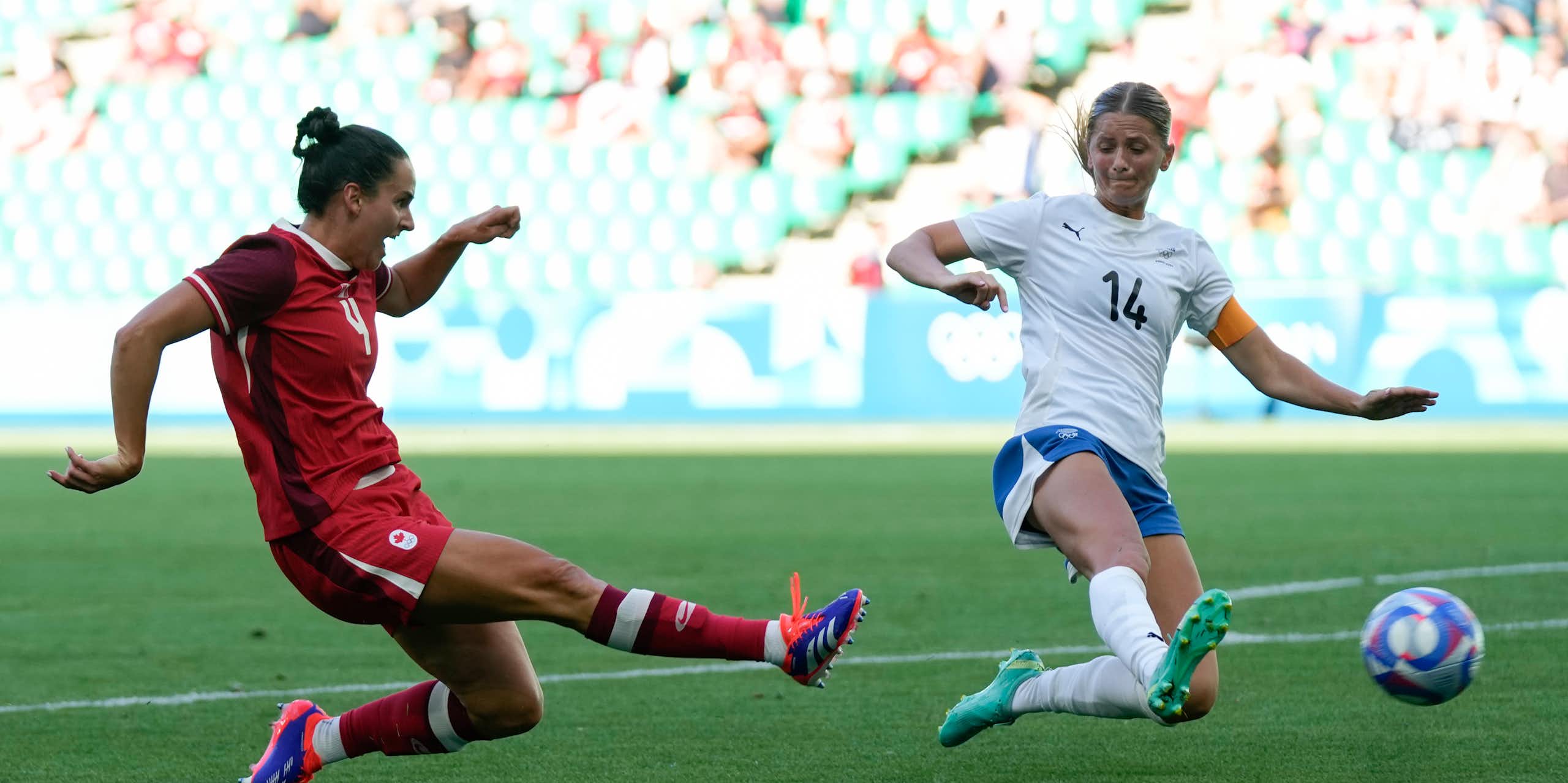 A woman in a red jersey kicks a ball while another in white attempts to deflect it