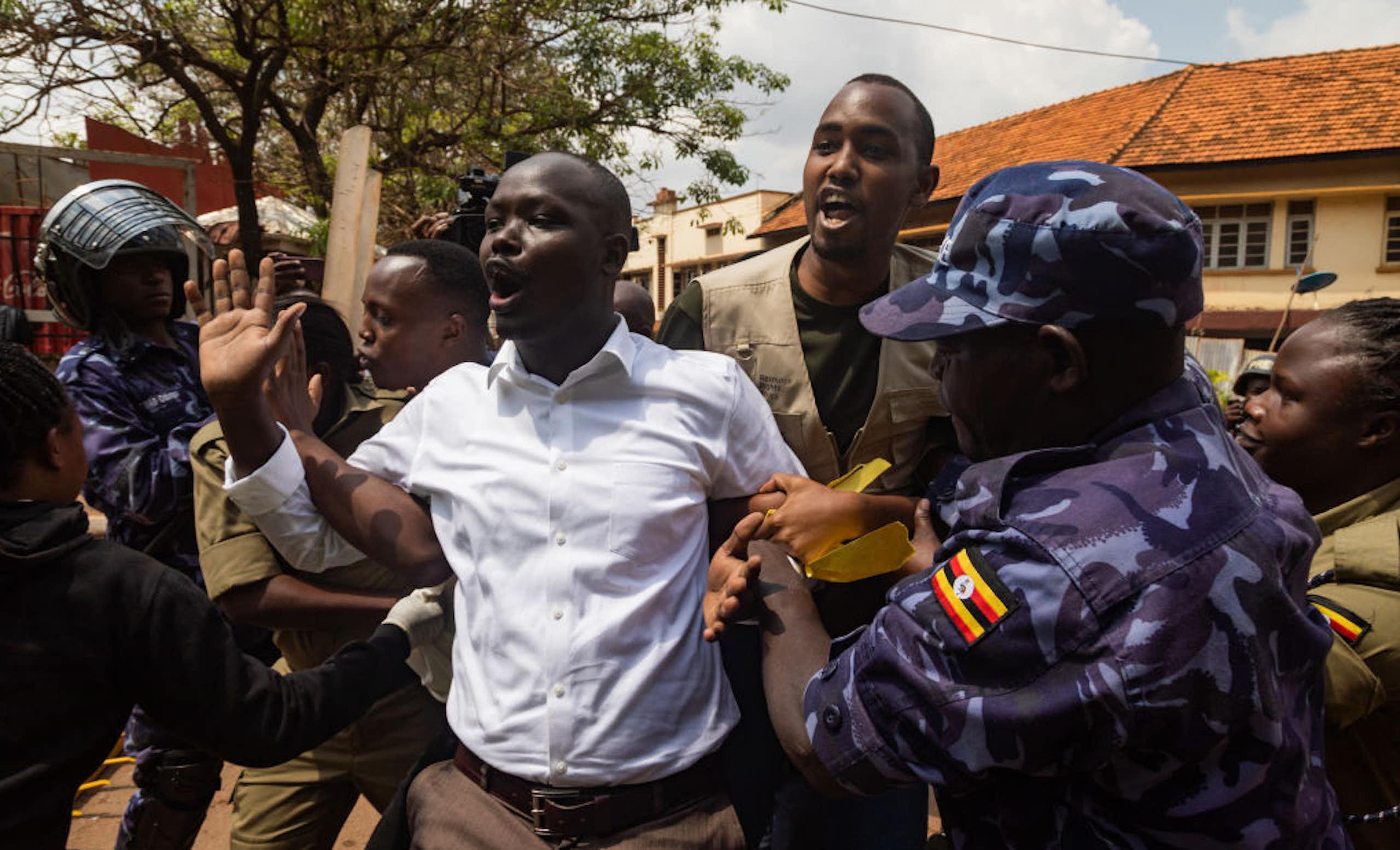 People in camouflage uniform and a brown uniform hold the arms of a man in civilian clothes