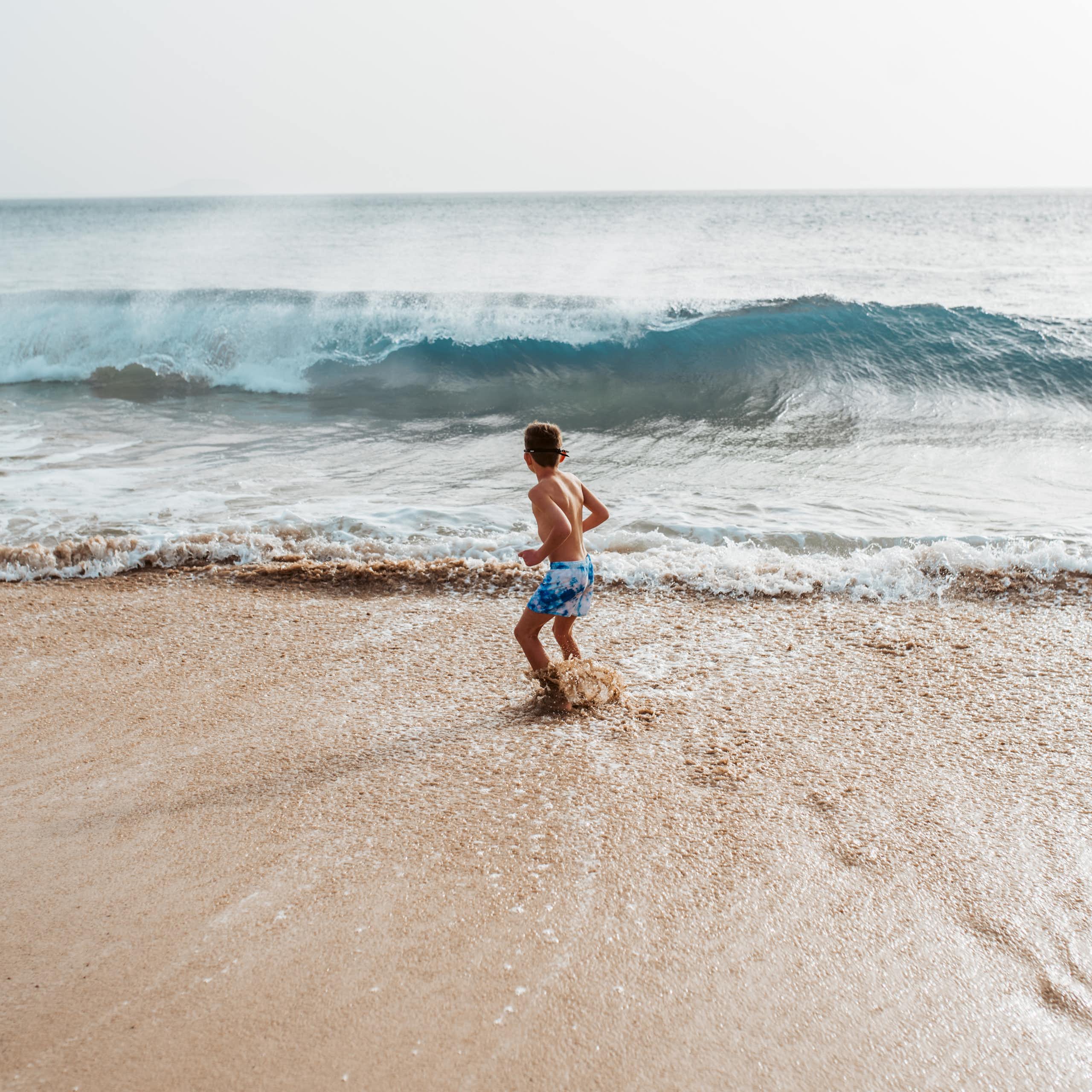 Un enfant hésite à se jeter dans les vagues, sur une plage face à la mer.