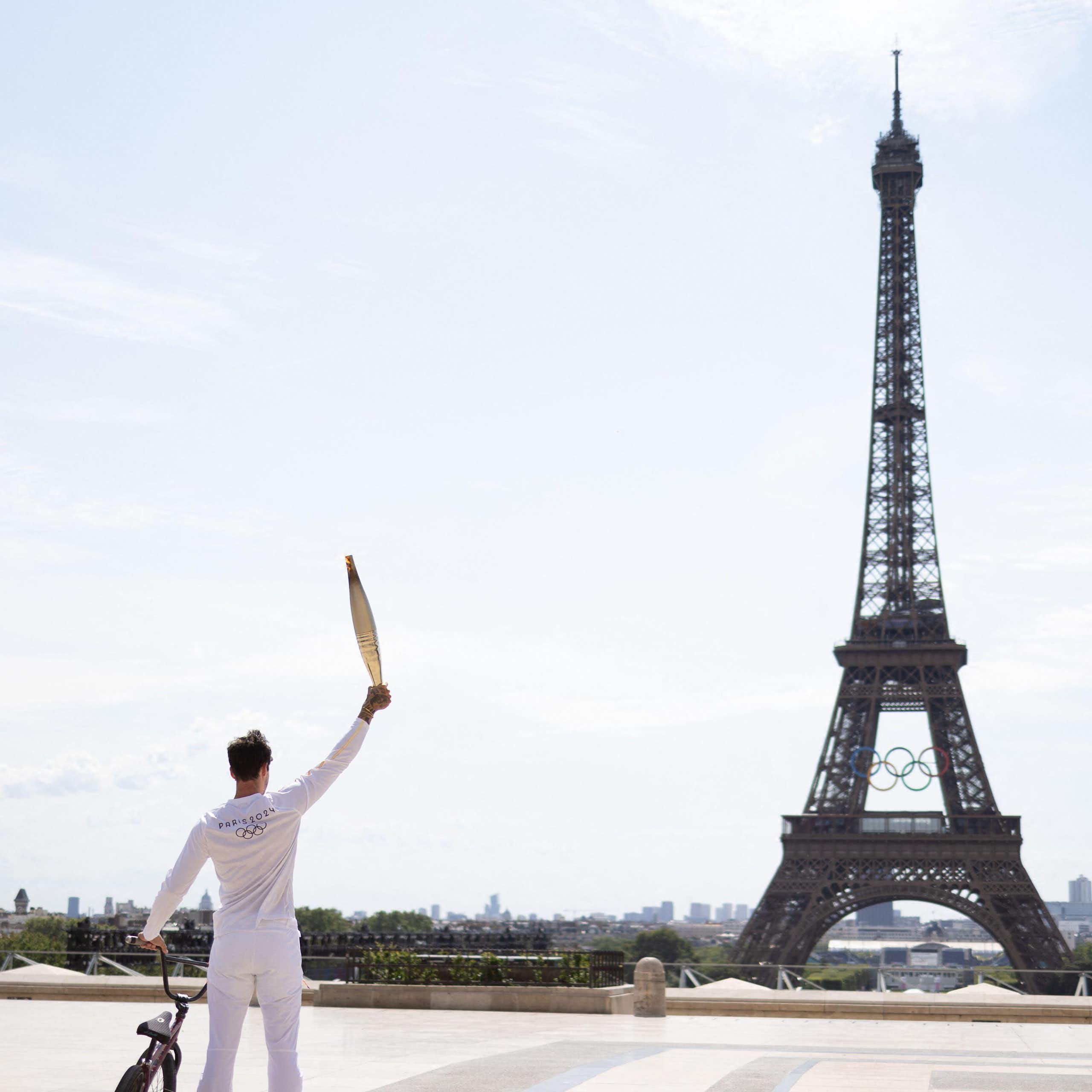 A man standing next to his BMX bike holding the Olympic torch aloft in front of the Eiffel Tower.
