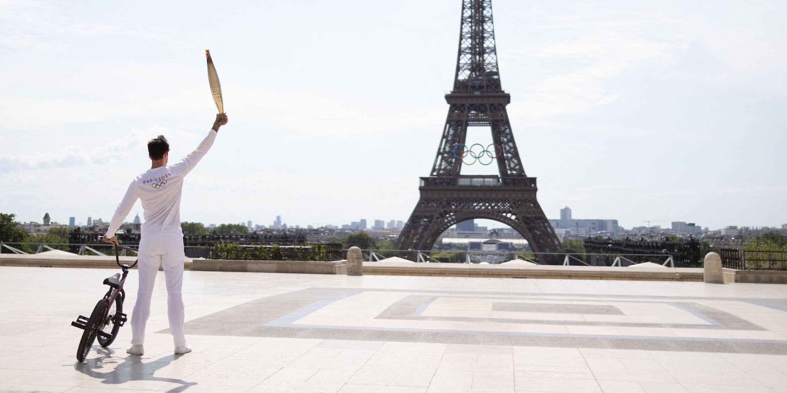 A man standing next to his BMX bike holding the Olympic torch aloft in front of the Eiffel Tower.