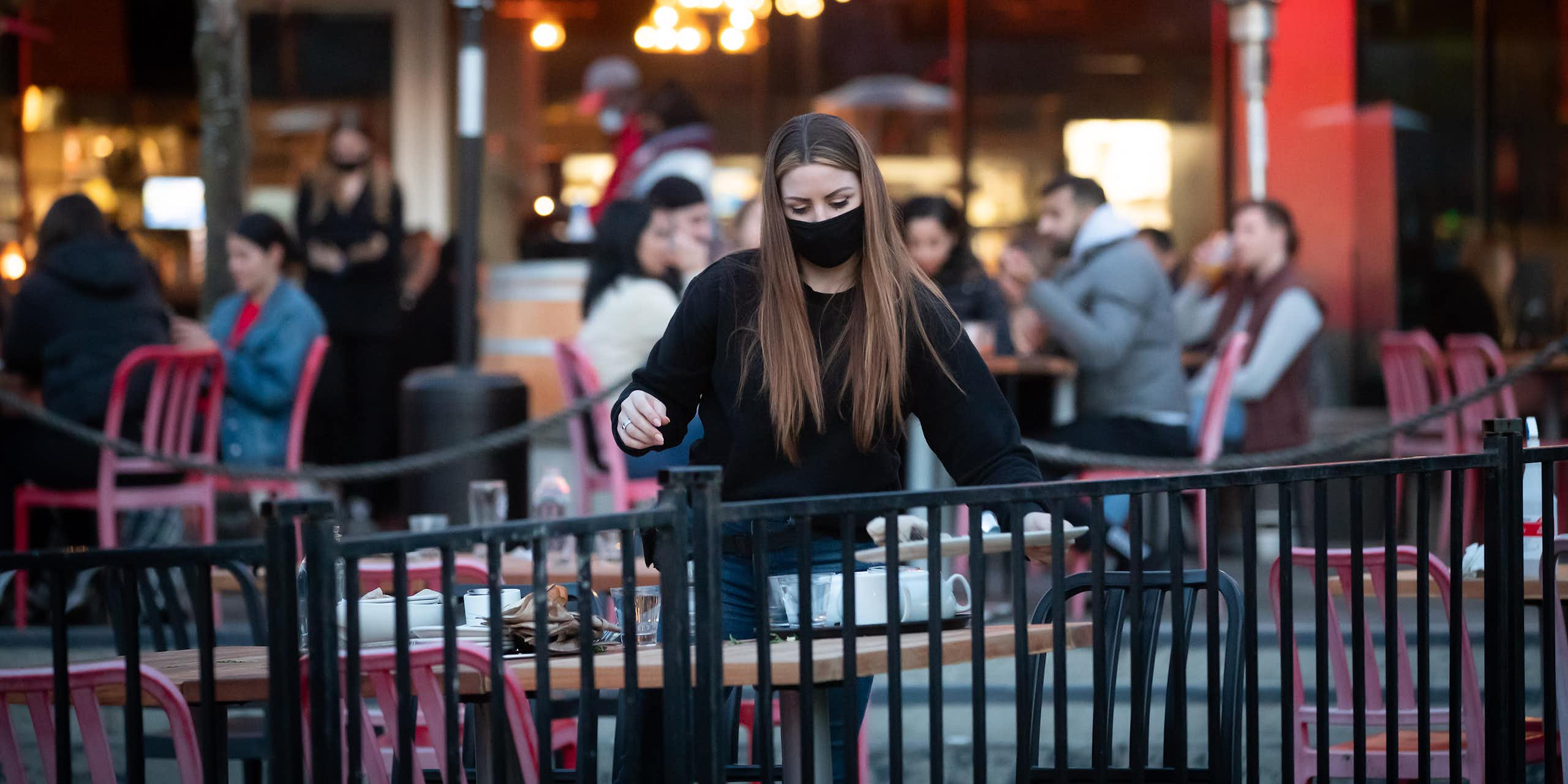 A young woman in a face mask clears dishes from an outdoor patio table in front of a restaurant