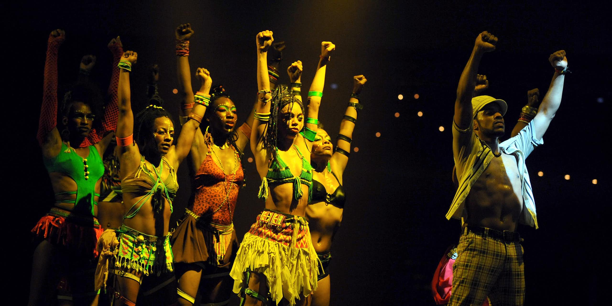 A stage-lit setting where a man with his shirt unbuttoned stands with both fists in the air. Behind him five women emulate his pose.