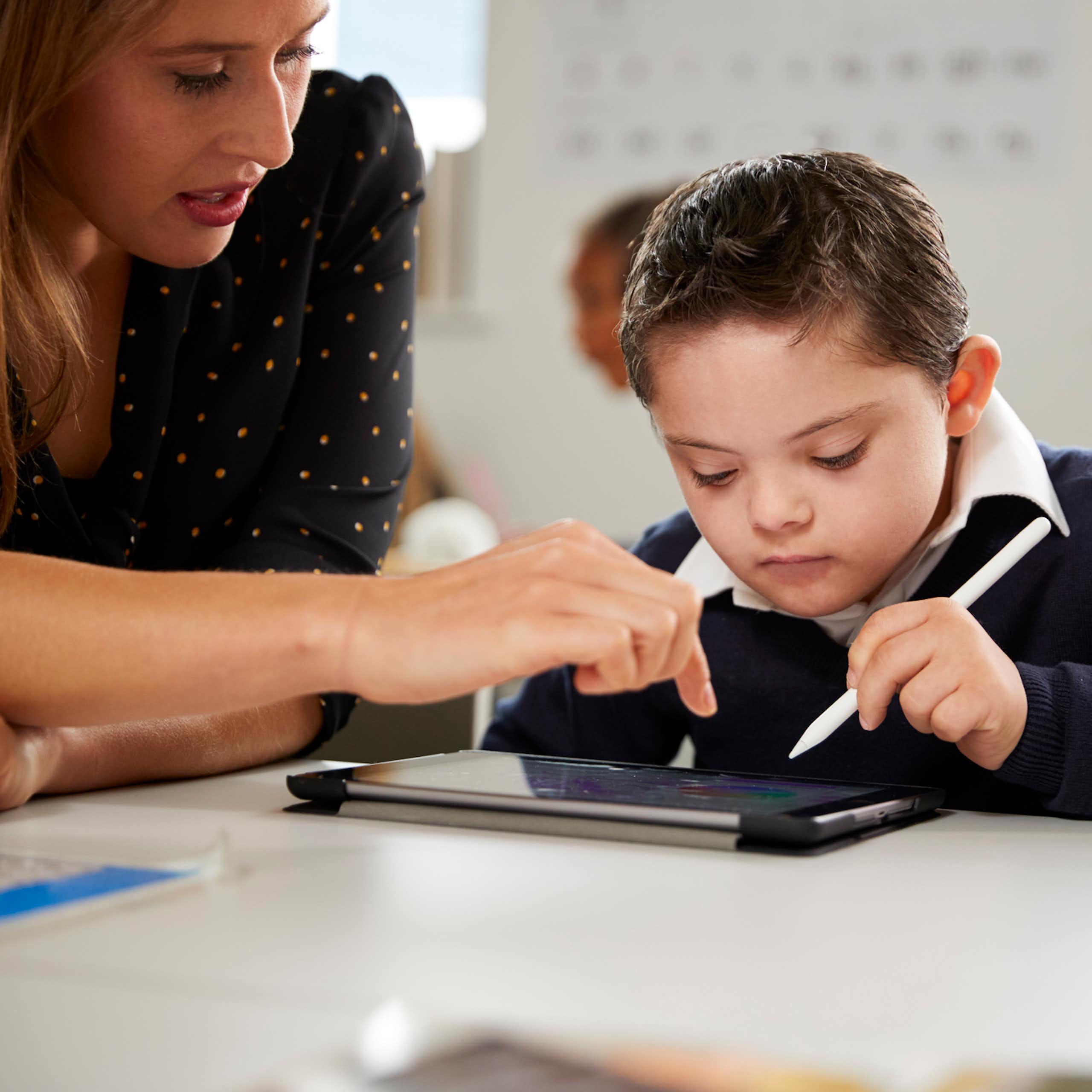 A woman leans over a desk. A young boy uses a stylus to work on a tablet. 