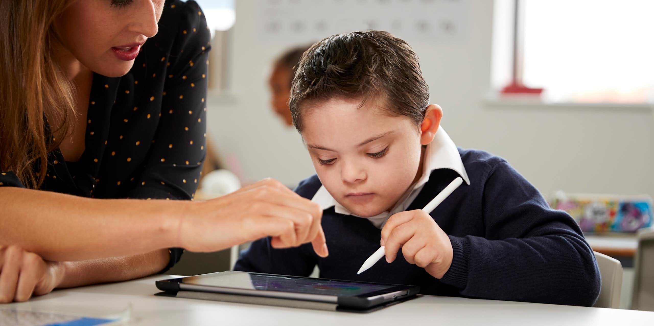 A woman leans over a desk. A young boy uses a stylus to work on a tablet. 