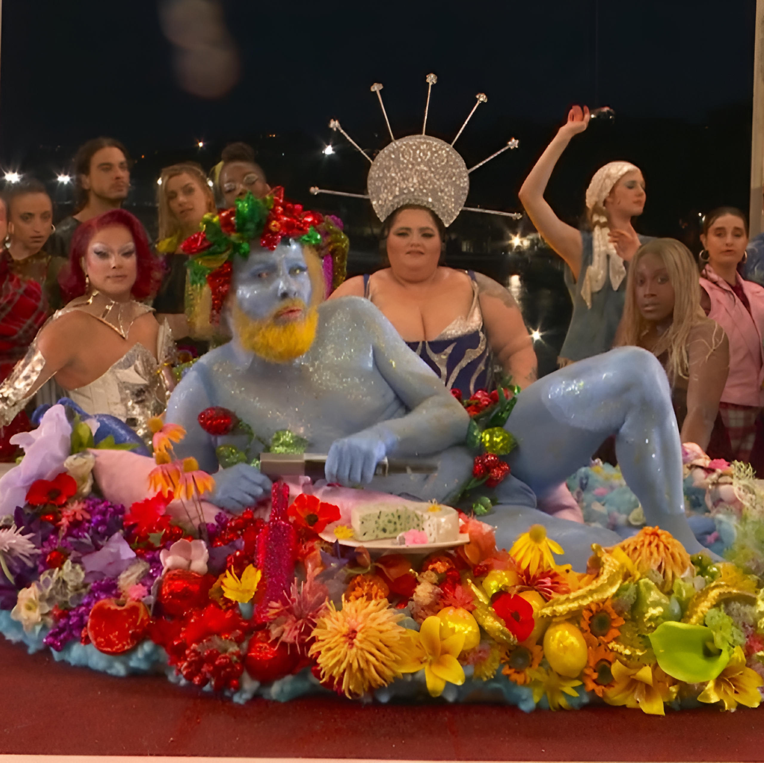 Un homme peint en bleu au front ceint d'une couronne de fleurs allongé sur une table couverte de fleurs ; derrière la table, une dizaine de personnes habillées de façon extravagante